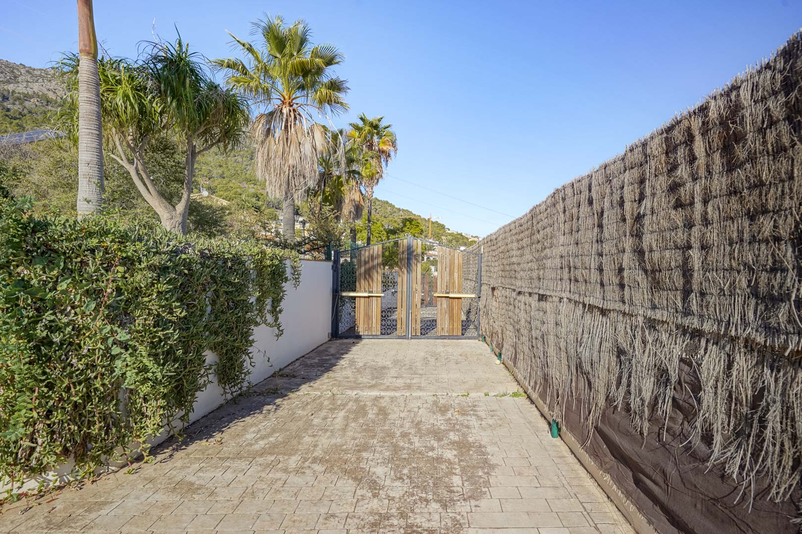 Ingang van het perceel met een bestrate oprit en houten poort, geflankeerd door mediterrane beplanting, palmbomen en een natuurlijke heidemat omheining.