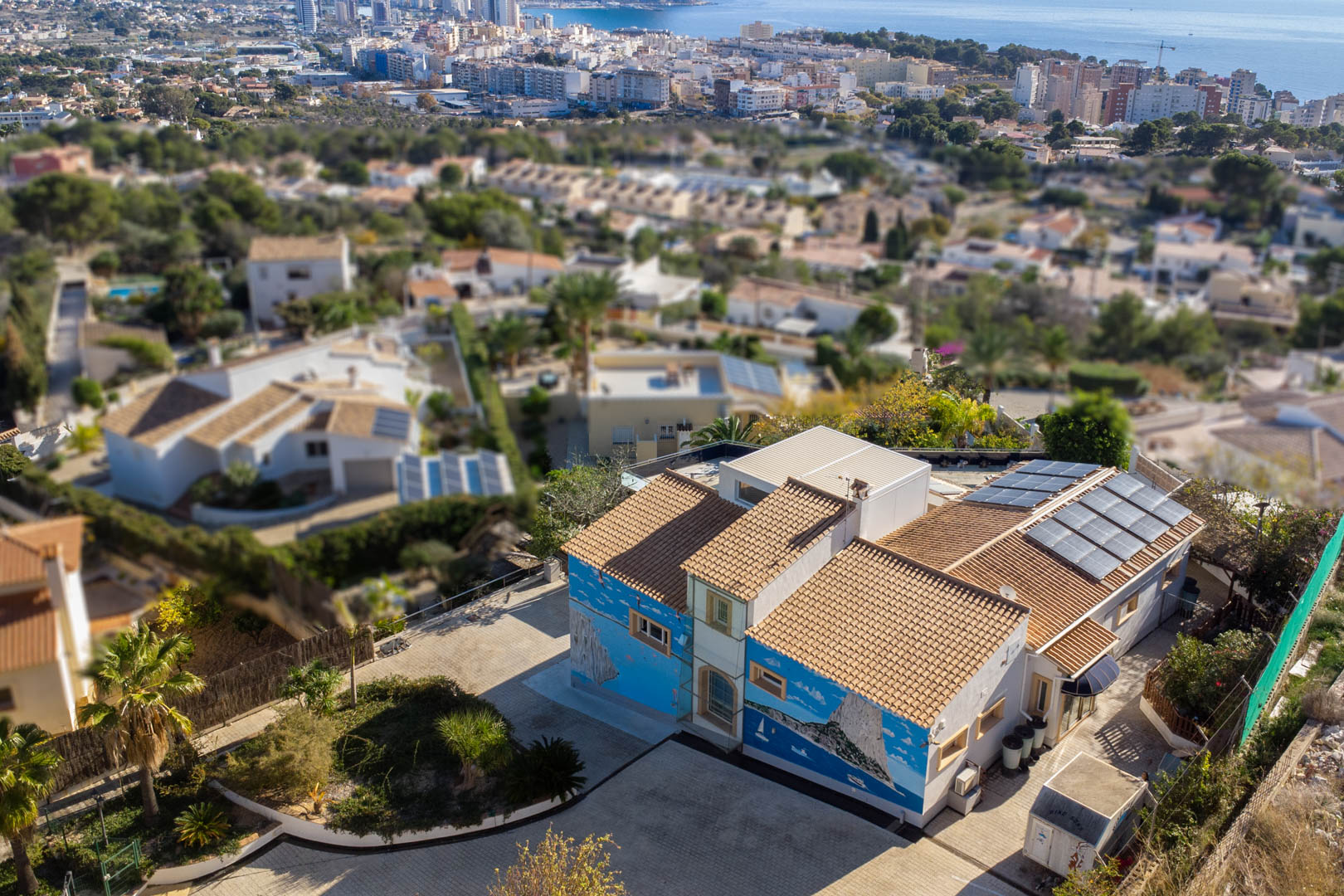 Luchtfoto van de villa met artistieke muurschilderingen, pannendak en zonnepanelen, gelegen in een woonwijk met uitzicht op de kustlijn en skyline van Calpe.