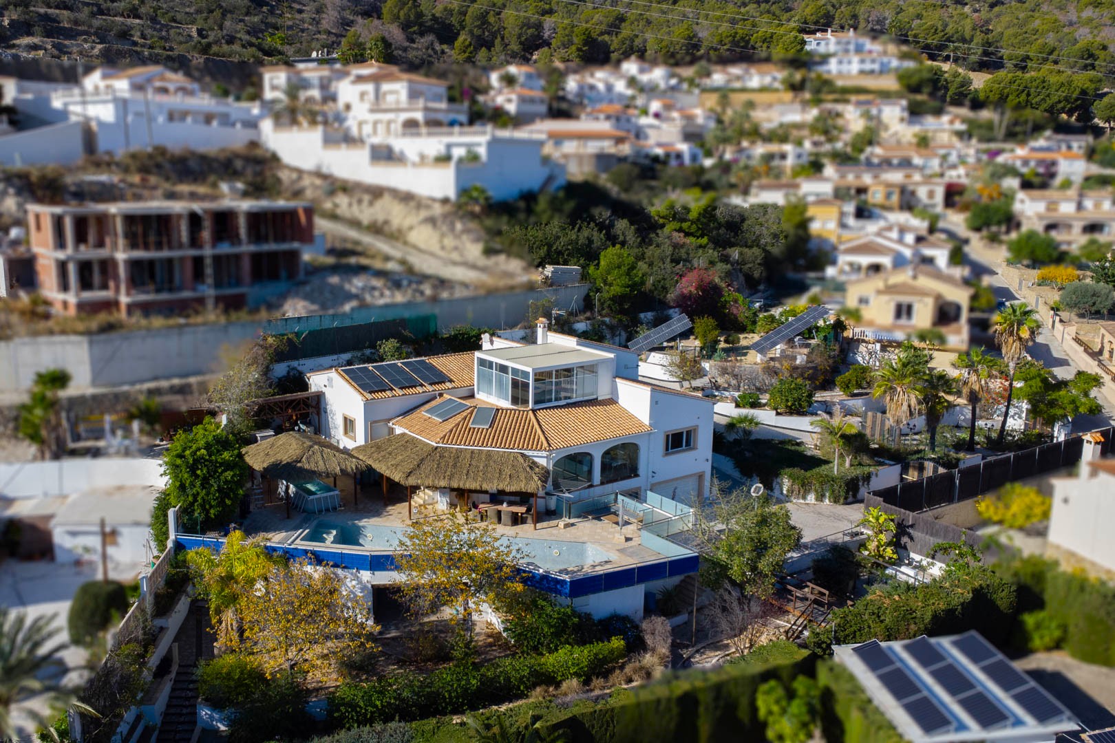 Luchtfoto van de villa met het zwembad, rieten pergola's, zonnepanelen op het dak en gemotoriseerde zonnetrackers in de aangelegde tuin.