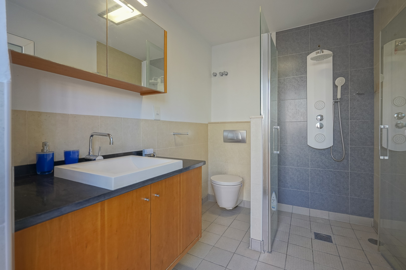 Moderne badkamer met houten wastafelmeubel, donker blad en inloopdouche. Voorzien van een hydromassagepaneel, glazen wand en een combinatie van beige en grijze tegels.