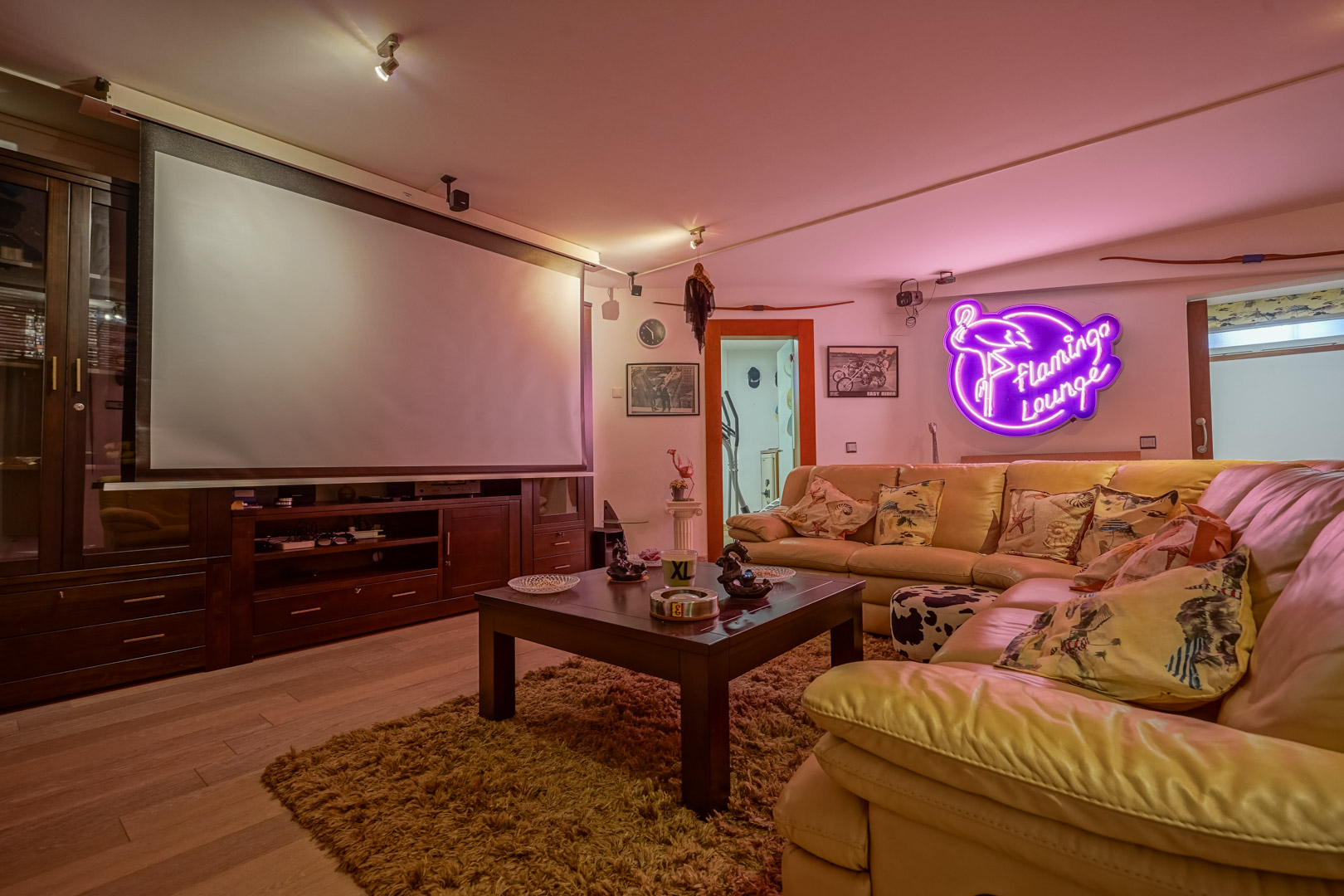 Multimediakamer met projectiescherm, donkerhouten kastenwand en een lederen hoekbank. De ruimte is voorzien van neon sfeerverlichting en een houten vloer.
