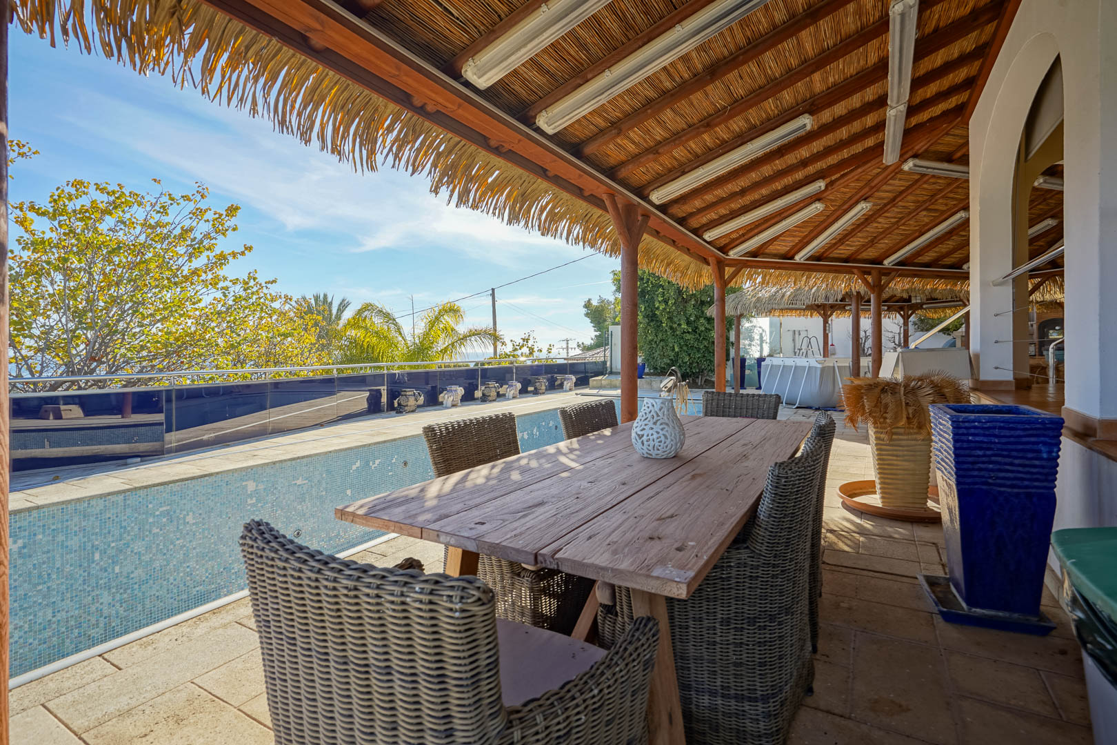 Overdekt terras met een rustieke houten eettafel en rieten stoelen onder een rieten pergola, grenzend aan het zwembad met uitzicht op de tuin.
