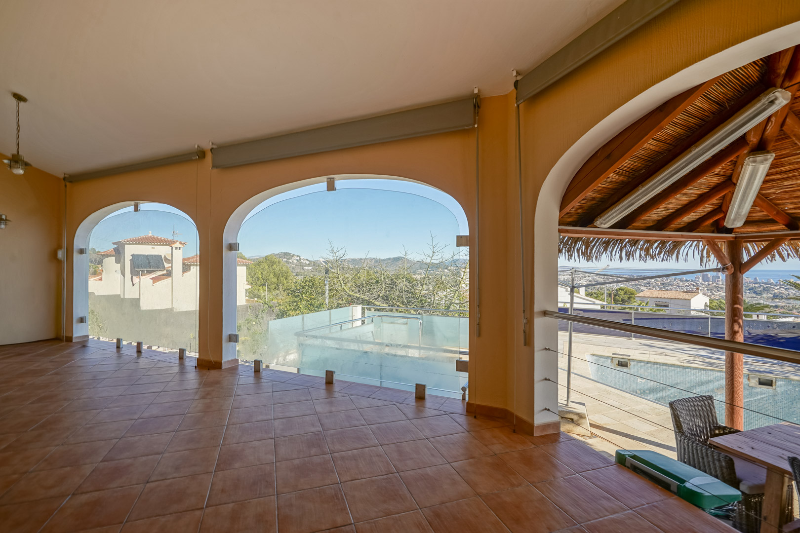 Overdekt terras met terracotta tegelvloer en glazen boogopeningen met uitzicht op het zwembadgedeelte en de kustlijn van Calpe.