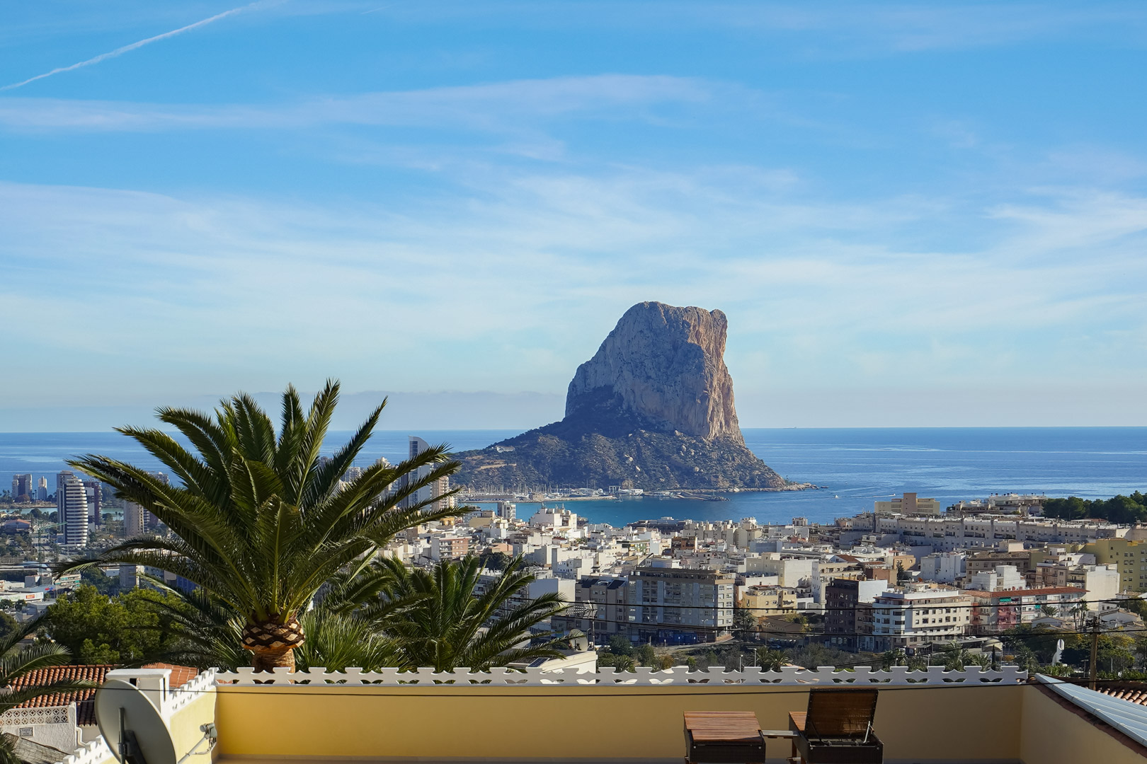 Panoramisch uitzicht vanaf het terras op de Peñón de Ifach en de skyline van Calpe, met de Middellandse Zee op de achtergrond en houten ligstoelen.
