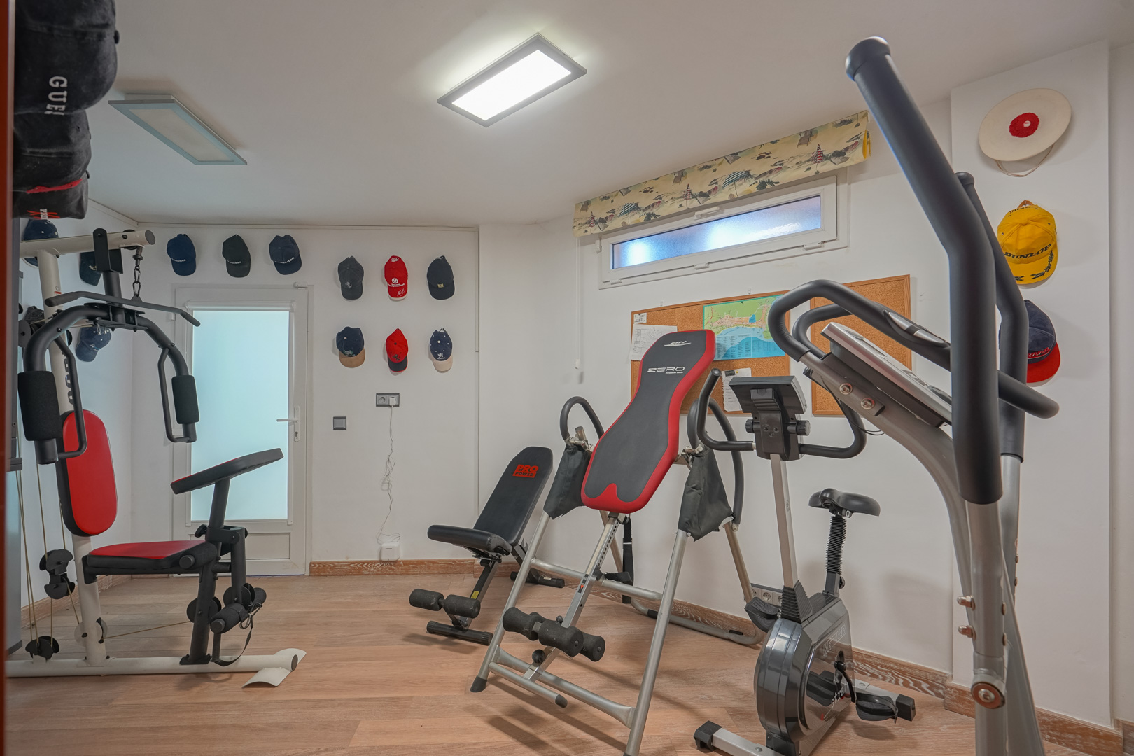 Privé-fitnessruimte met houten vloer, uitgerust met een multistation, hometrainer en inversiebank onder heldere led-paneelverlichting.
