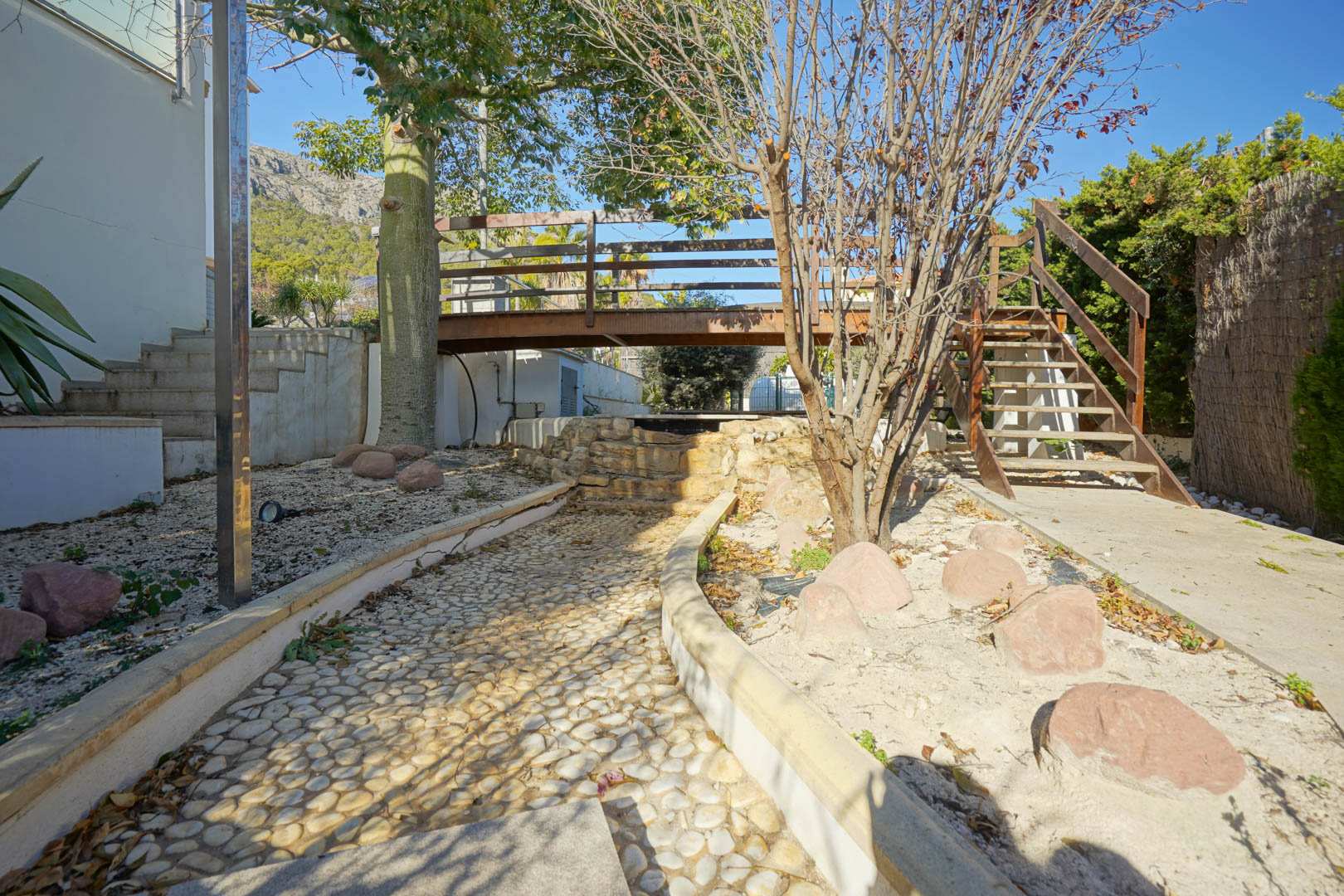Tuin met een houten loopbrug over een decoratieve stenen waterval, een geplaveid kiezelpad en houten trappen met uitzicht op de Sierra de Oltamar.