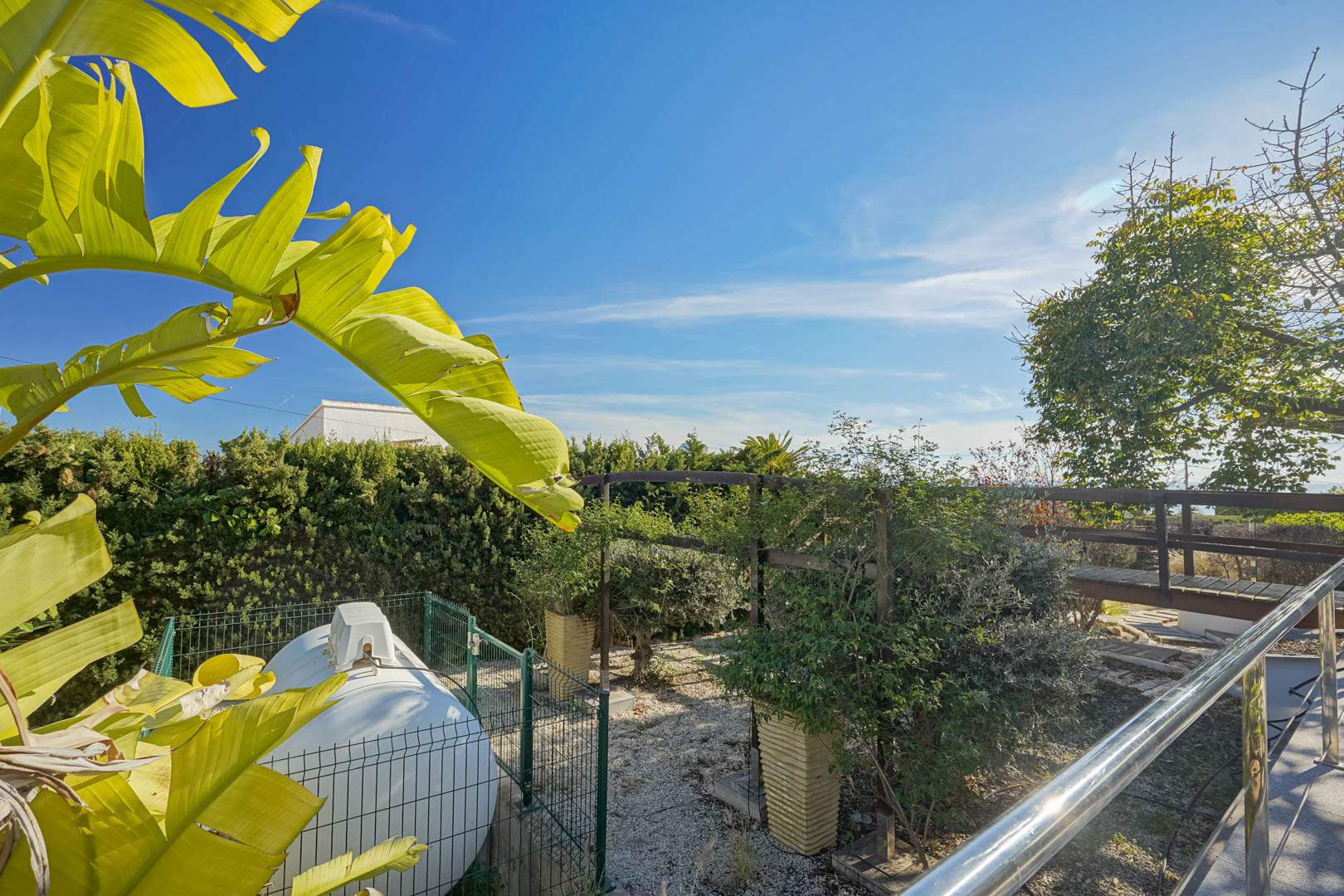 Zijtuin met een omheinde gastank, een houten loopbrug en tropische vegetatie onder een helderblauwe lucht.