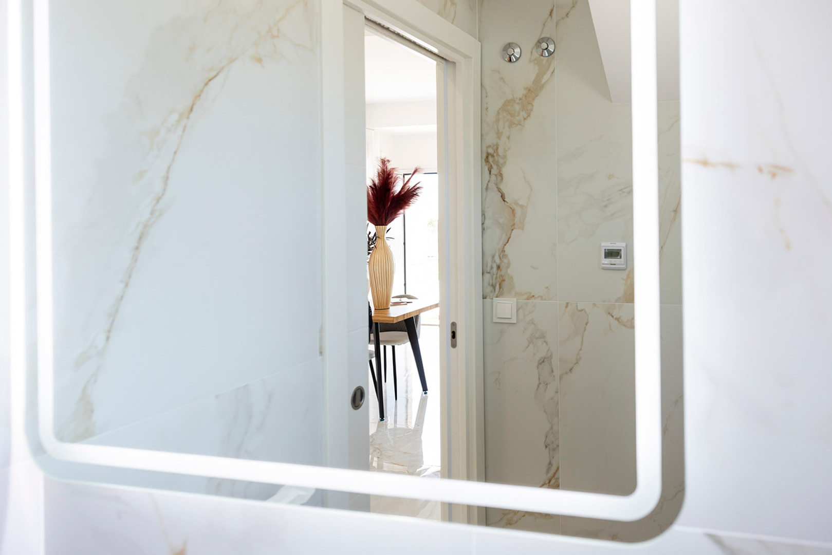Badkamer met wandtegels in marmerlook en een spiegel met LED-verlichting, met doorkijk naar de eethoek via een witte deuropening.