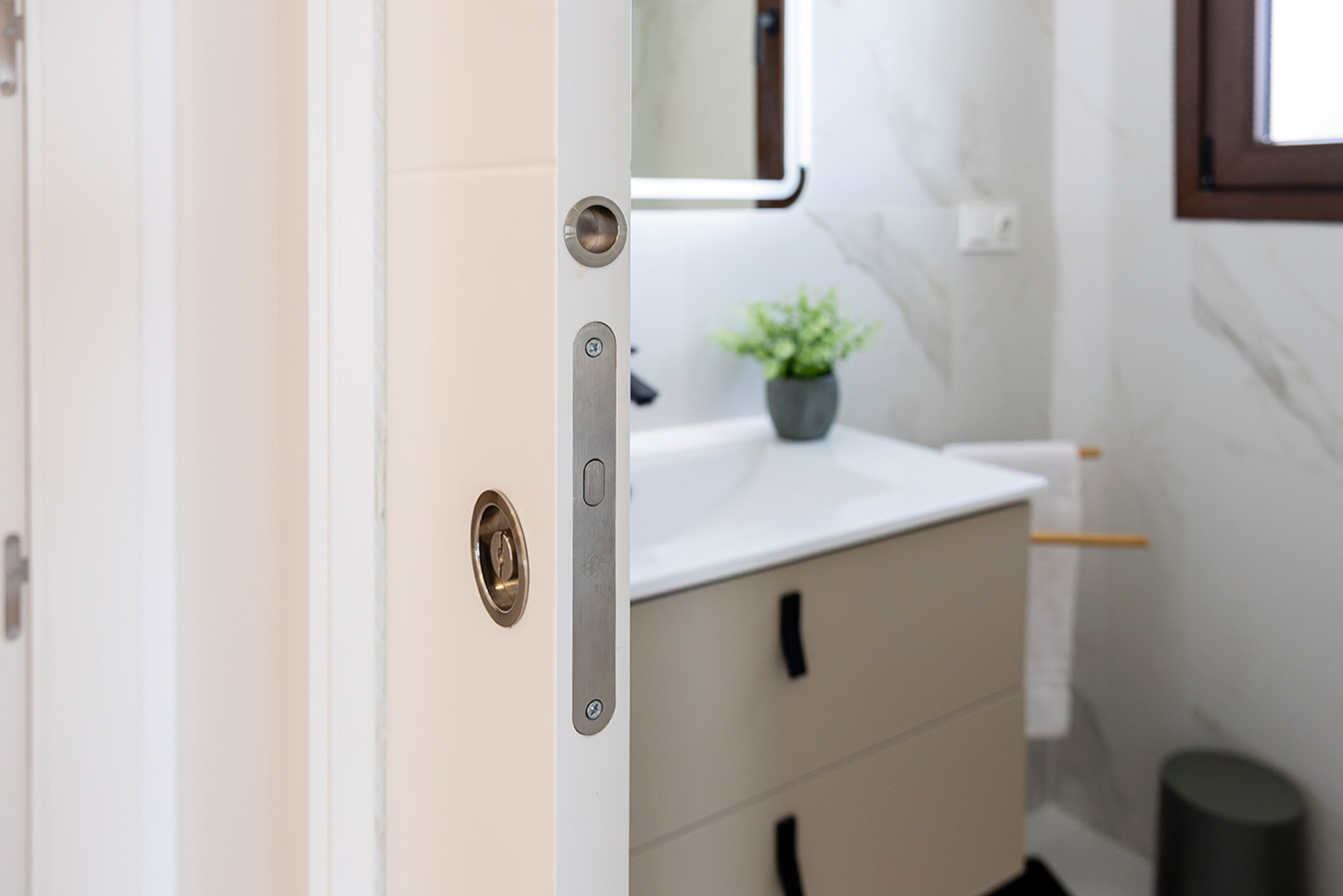 Detail van een witte schuifdeur met metalen inbouwbeslag, die toegang biedt tot een moderne badkamer met marmerlook tegels en een eigentijds wastafelmeubel.