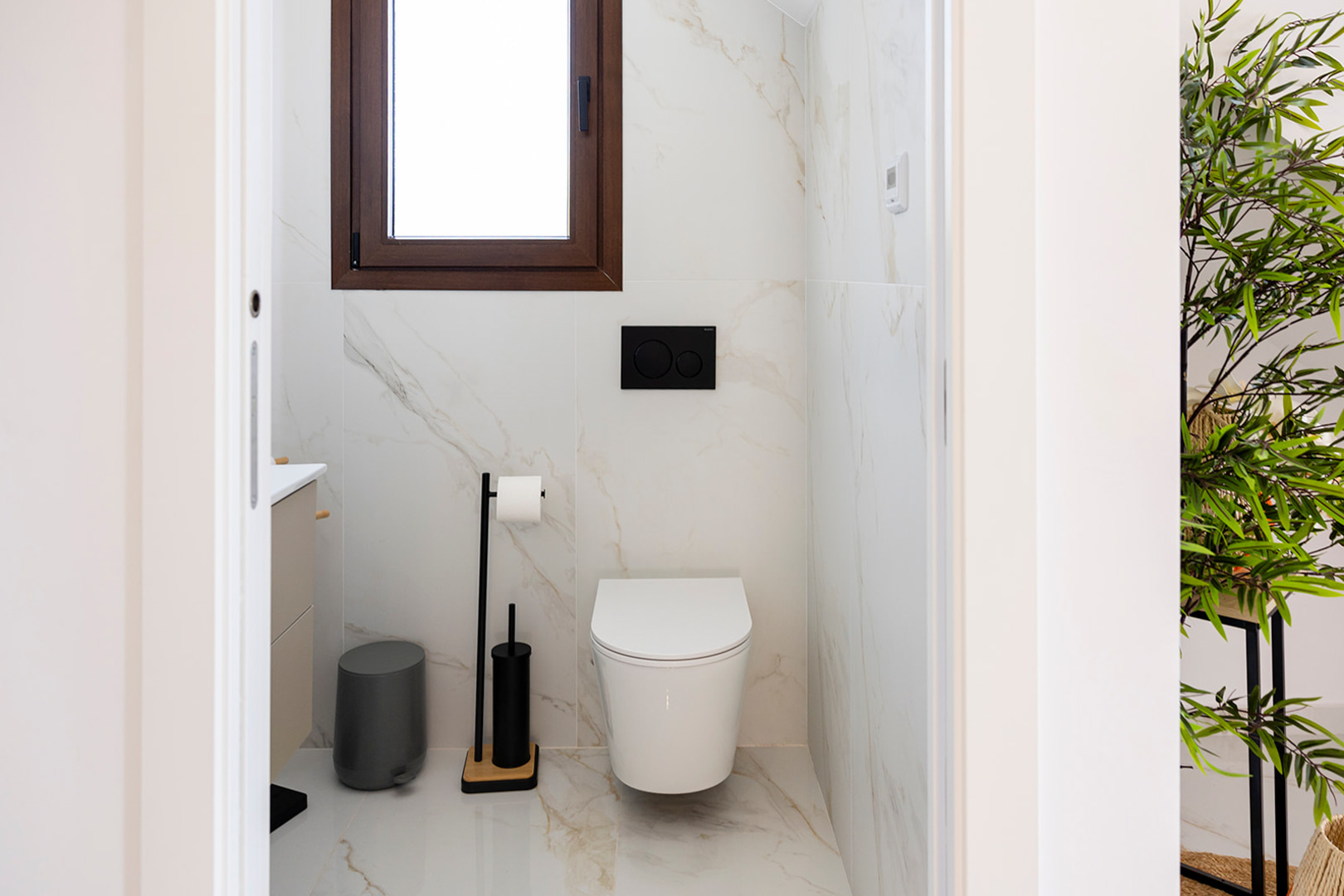Gastentoilet met marmerlook tegels, een hangend toilet en matzwarte accessoires. Het raam met houten kozijn zorgt voor natuurlijke lichtinval.