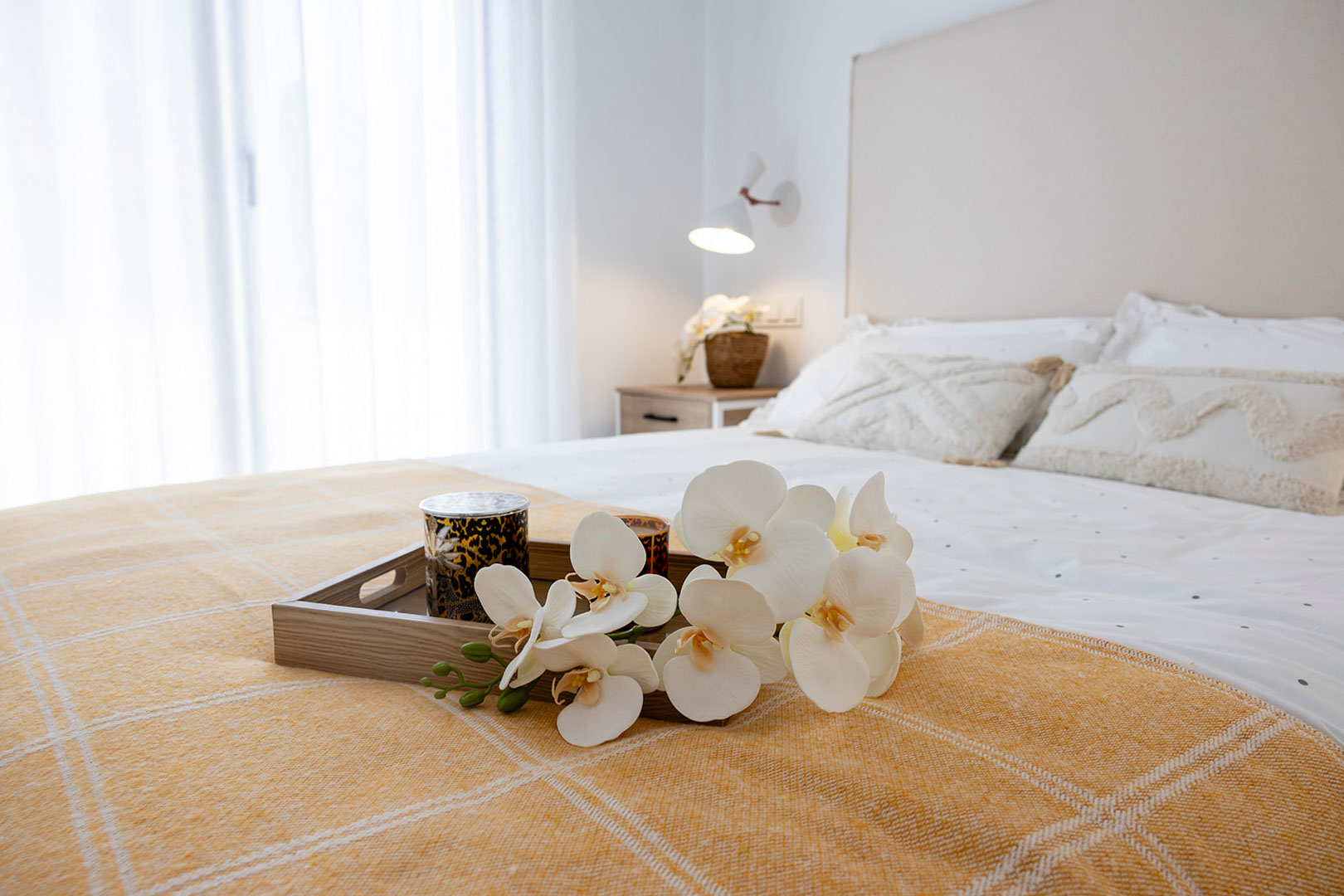 Lichte slaapkamer met gestoffeerd hoofdbord, wit en okergeel textiel, en een decoratief dienblad met orchideeën. Natuurlijk licht valt binnen door grote ramen met vitrages.