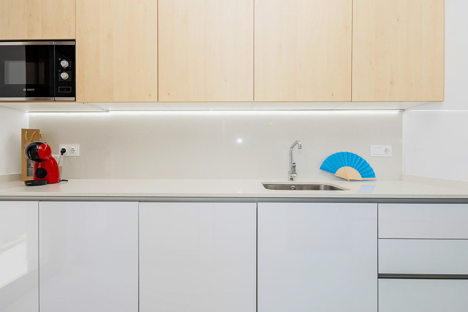Moderne keuken met bovenkasten in licht hout, witte onderkasten, een quartz werkblad en geïntegreerde LED-verlichting onder de kasten.