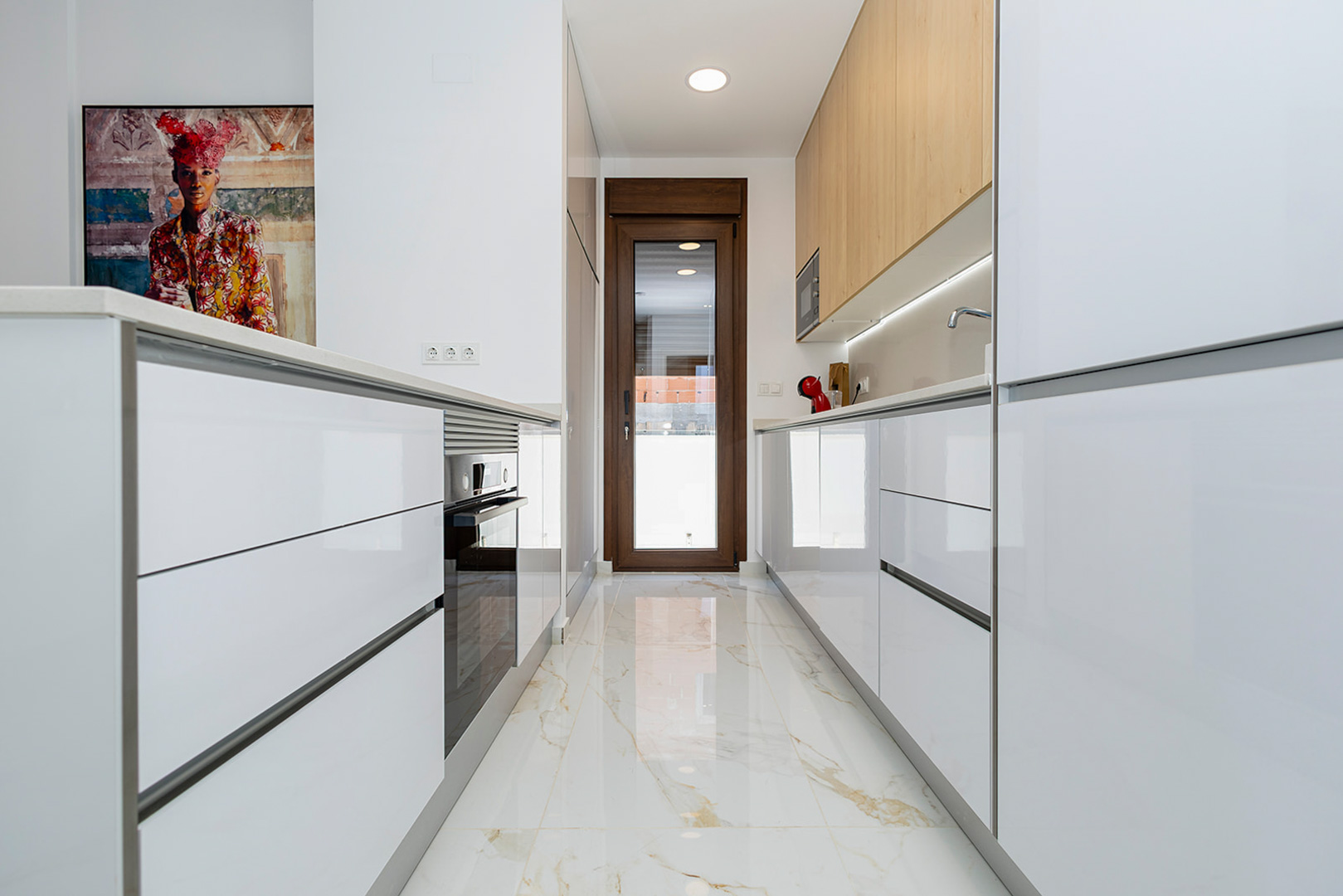 Moderne keuken met hoogglans witte kasten, houten bovenkasten en een porseleinen vloer met marmereffect. Voorzien van inbouwapparatuur en directe toegang tot buiten.