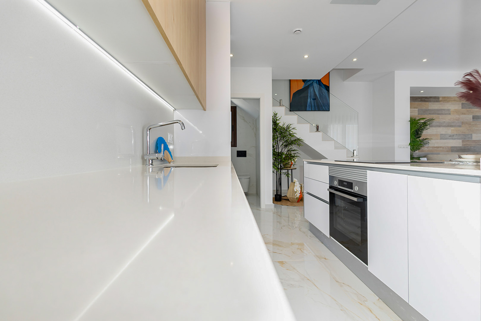 Moderne keuken met wit werkblad, LED-verlichting onder houten kasten en een kookeiland. Op de achtergrond is de trap met glazen balustrade en marmeren vloer te zien.