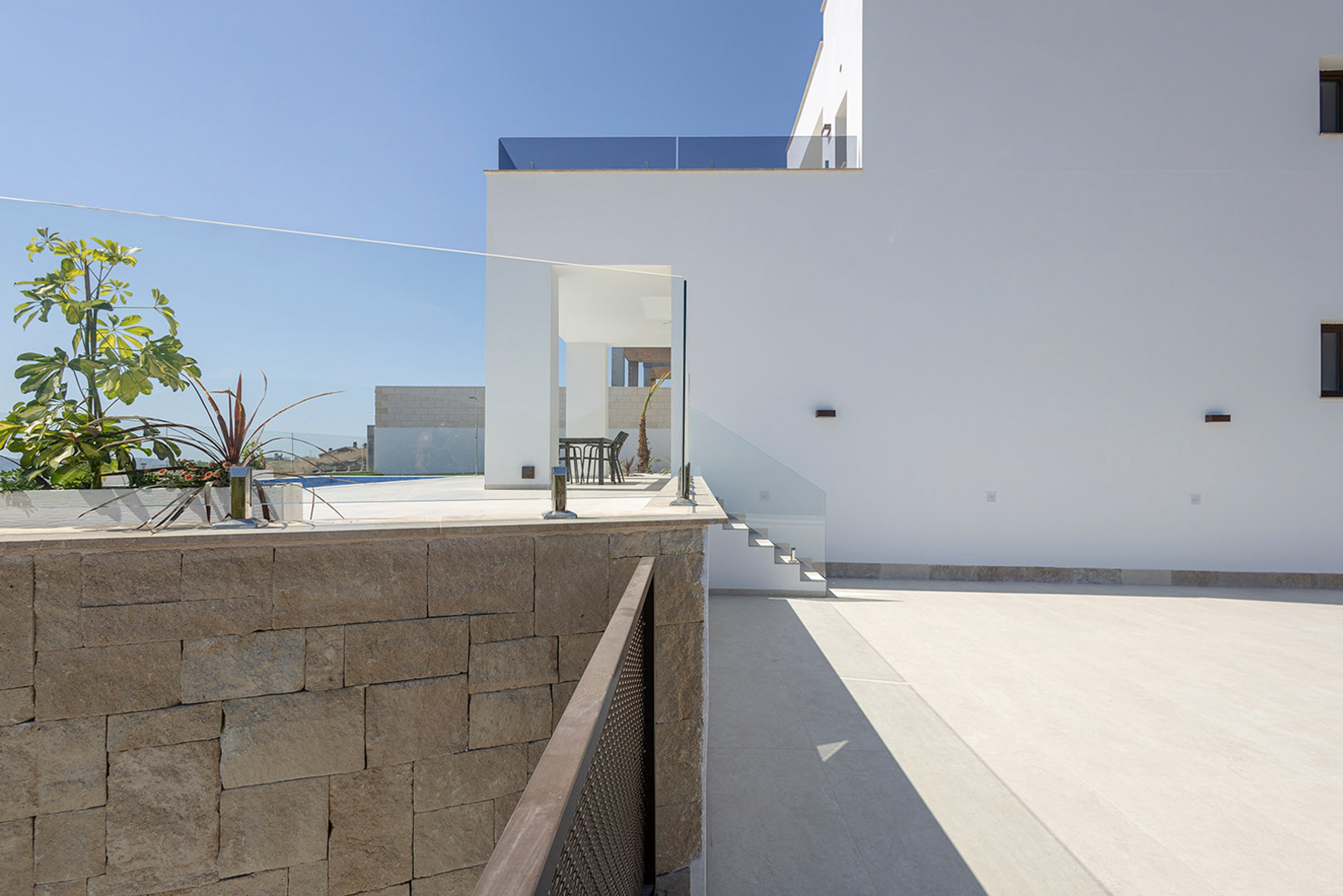 Moderne witte gevel met een natuurstenen muur, glazen balustrades en een geplaveide buitenruimte die toegang biedt tot het bovenliggende terras.