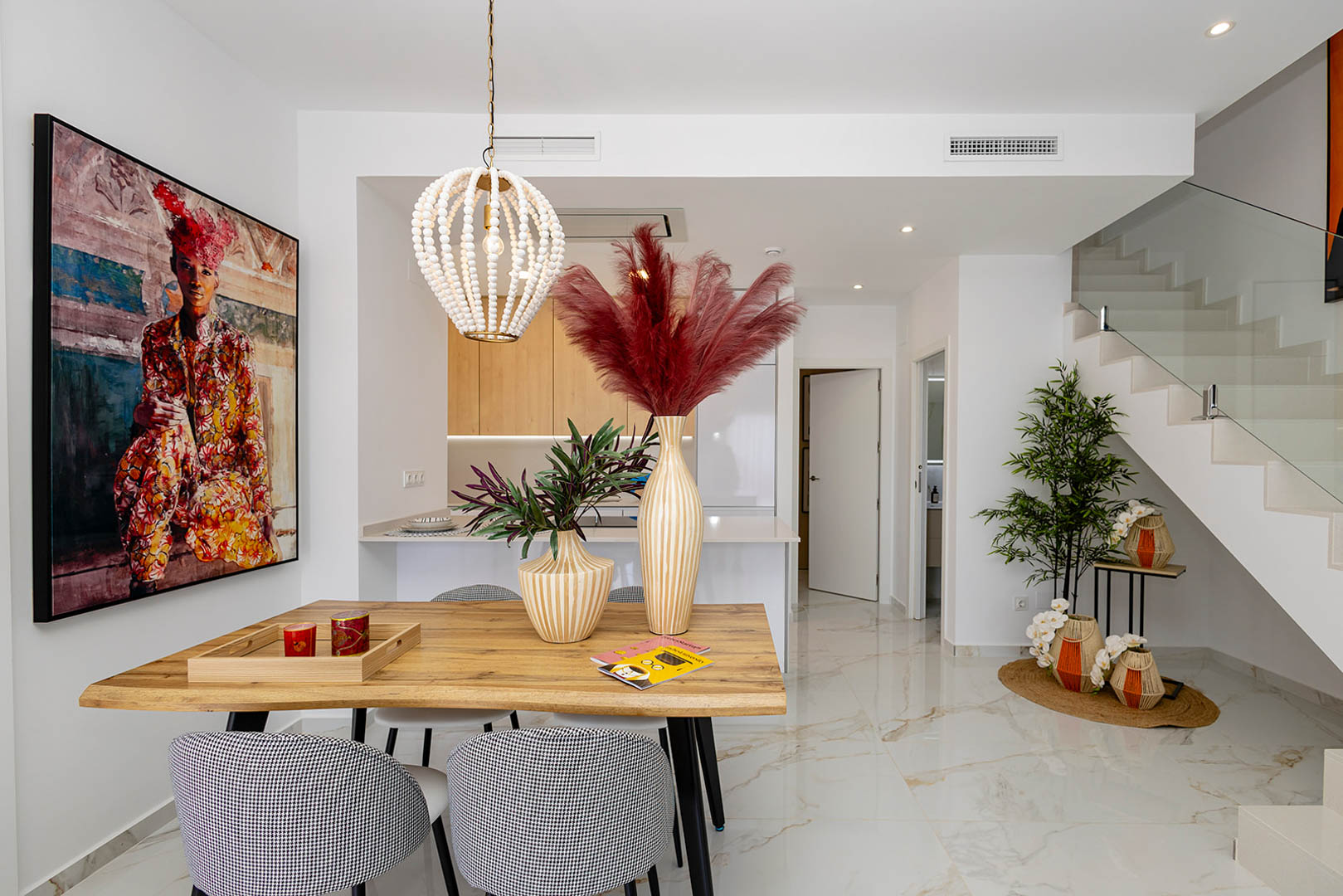 Open eetkamer met natuurhouten tafel, kralen hanglamp en marmerlook porseleinen vloer naast een moderne trap met glazen balustrade.