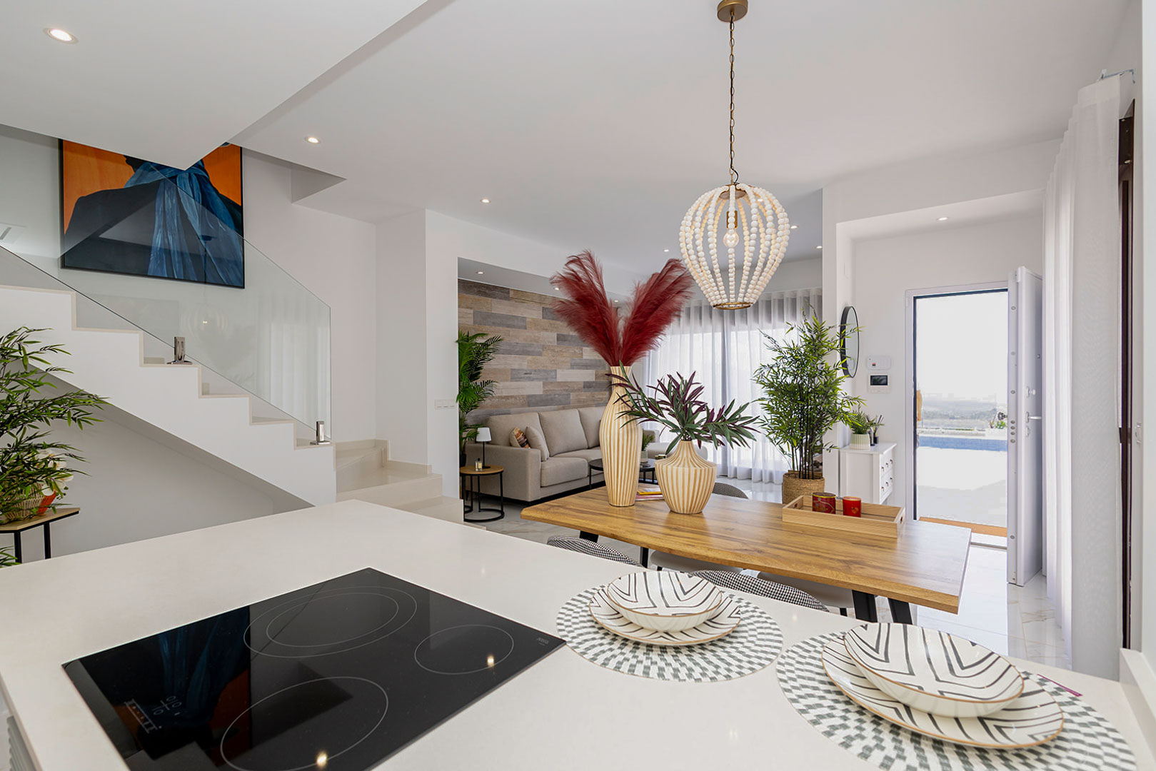Open woonruimte met kookeiland voorzien van inductiekookplaat, houten eettafel en een moderne trap met glazen balustrade.