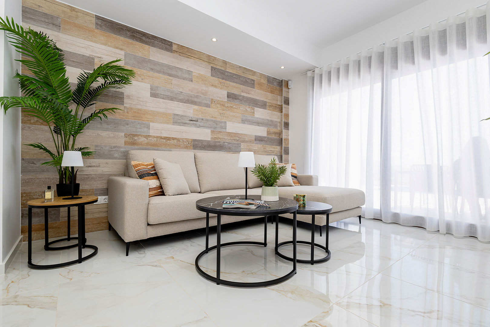 Woonkamer met een accentmuur van houten lamellen, gepolijste marmerlook vloertegels en een beige loungebank bij grote glazen schuifdeuren.
