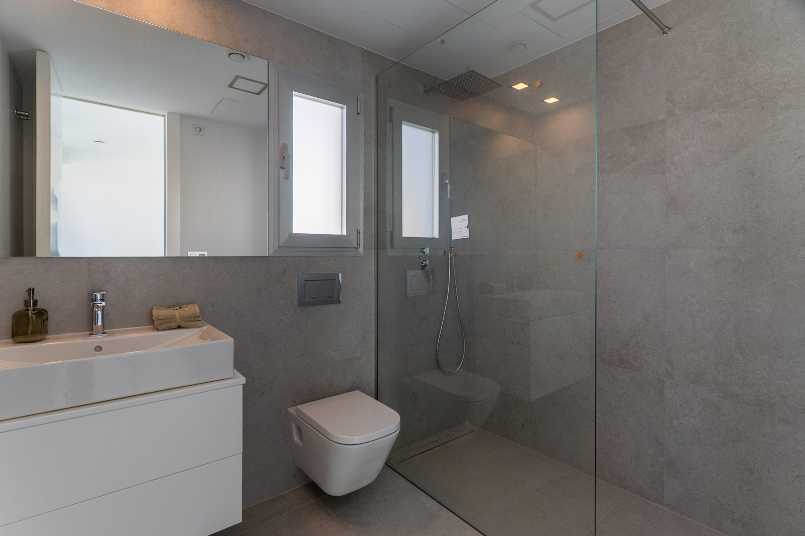 Eigentijdse badkamer met grijze tegels, inloopdouche met glazen wand, hangend toilet en geïntegreerde wastafel op een minimalistisch wit meubel.
