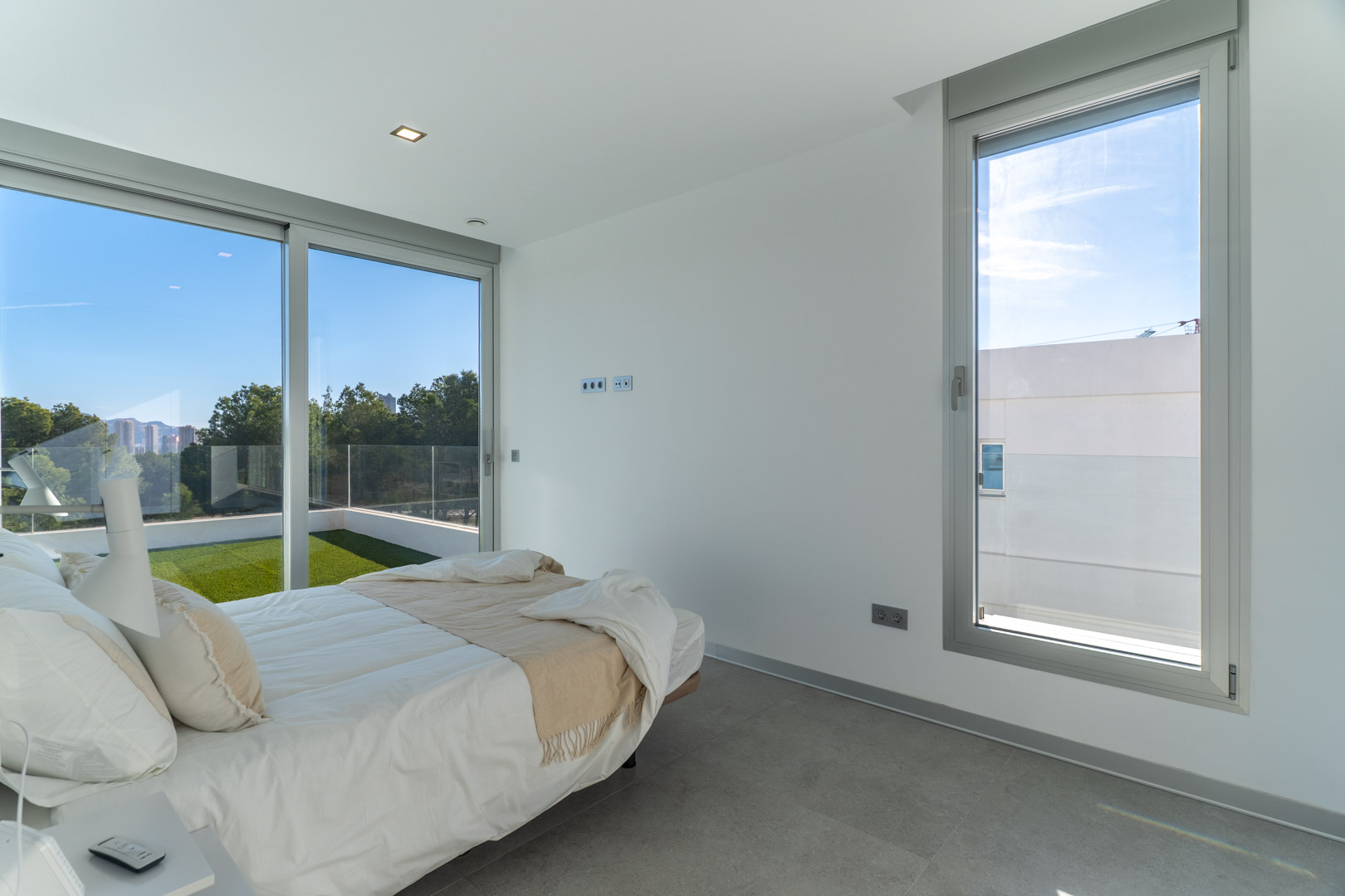 Lichte slaapkamer met grijze tegelvloer, grote aluminium schuifdeuren en directe toegang tot een privéterras met kunstgras en weids uitzicht.
