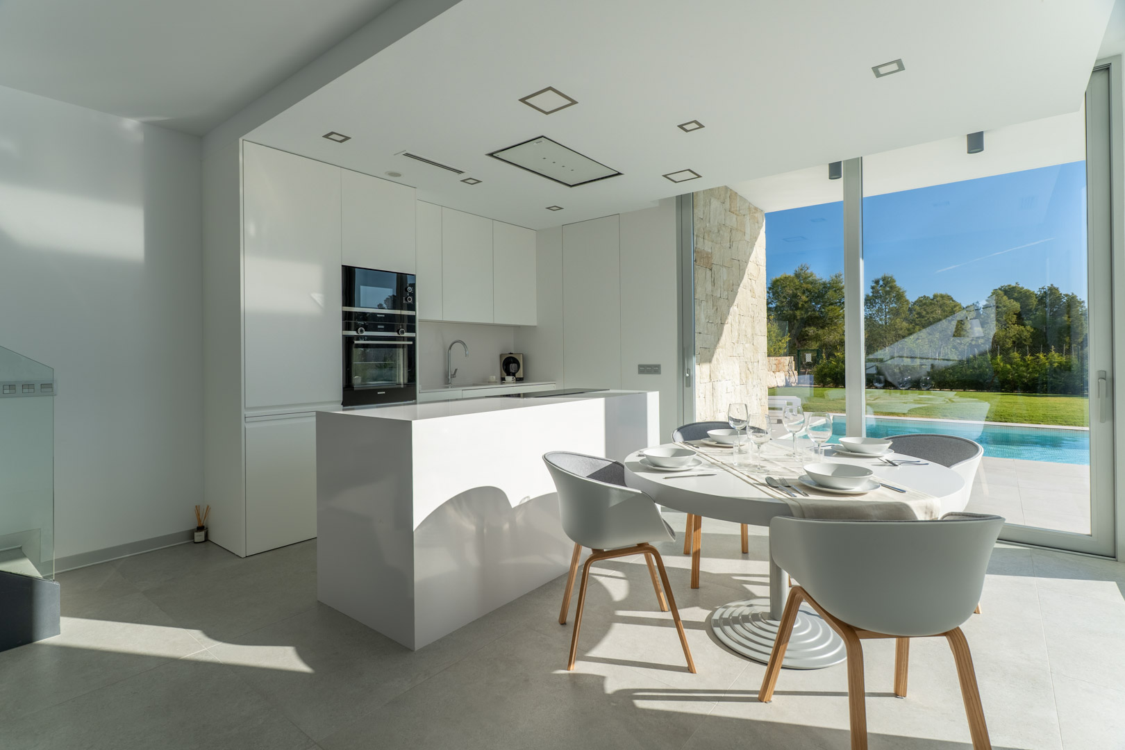 Minimalistische witte open keuken met kookeiland en eethoek, voorzien van kamerhoge ramen met uitzicht op het privézwembad en de tuin.