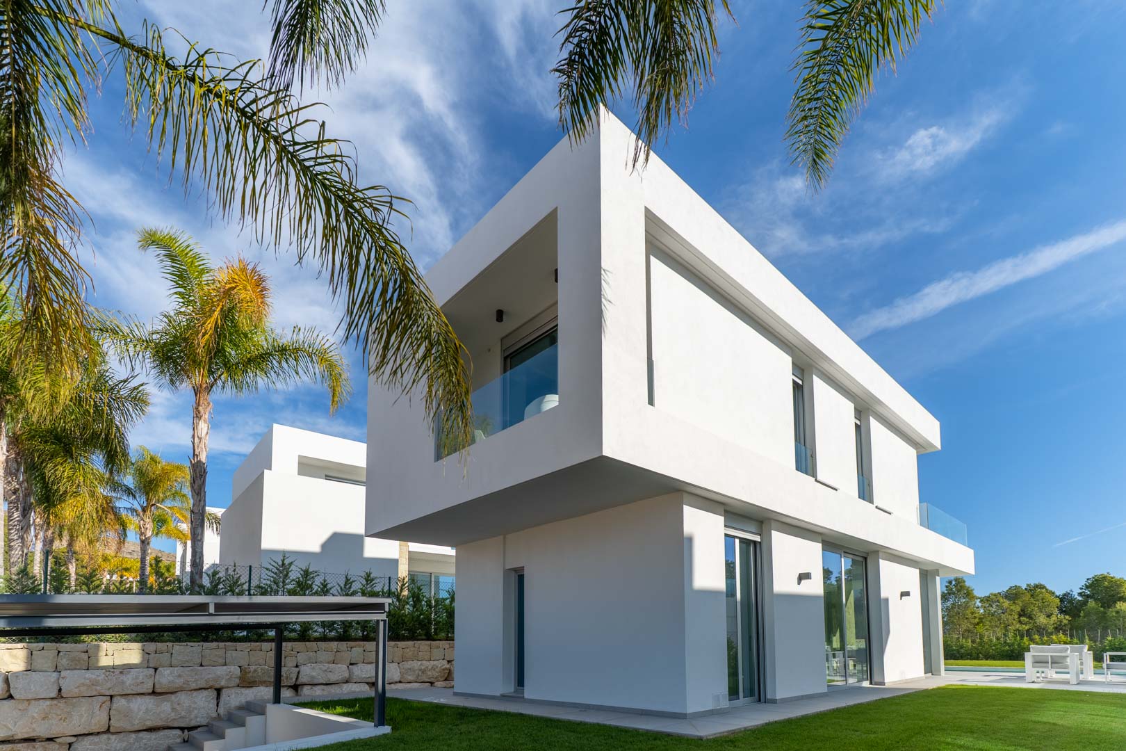 Moderne villa met twee verdiepingen, een overstekende bovenverdieping en kamerhoge glazen schuifdeuren. De tuin is voorzien van een gazon en karakteristieke palmbomen.