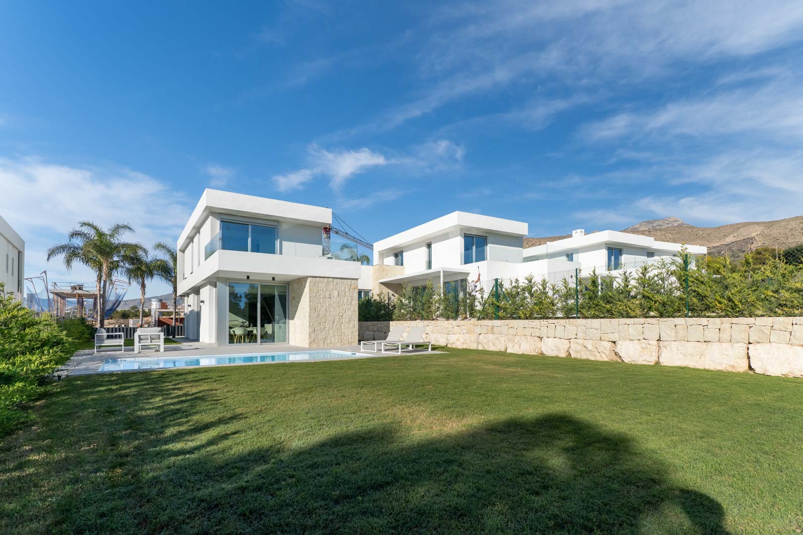 Moderne villa met witte gevel en natuurstenen details, privézwembad, groot gazon en uitzicht op de bergen onder een helderblauwe lucht.