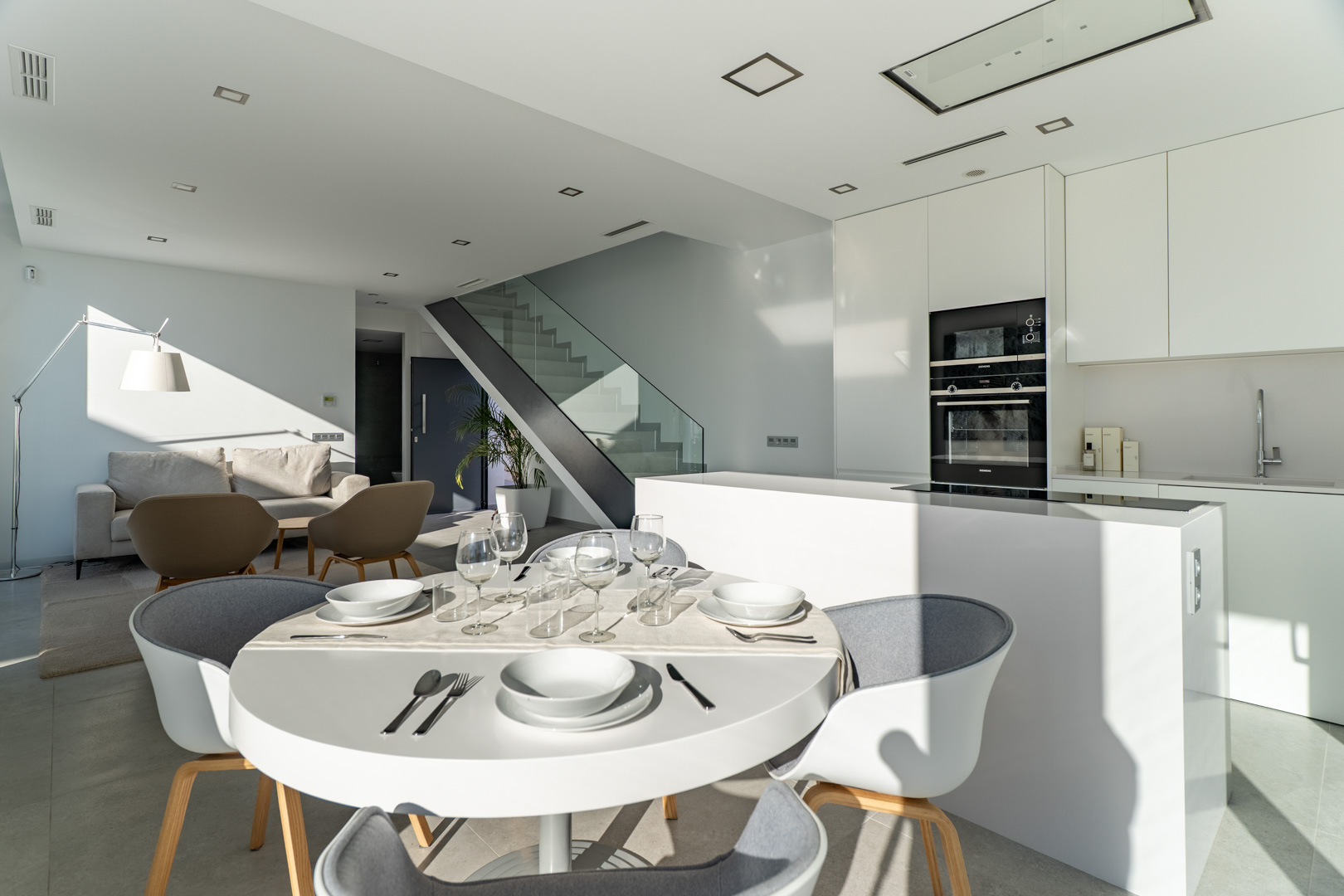 Open woonruimte met een minimalistische witte keuken, kookeiland en inbouwapparatuur. Een moderne trap met glazen balustrade vormt een centraal architectonisch element.
