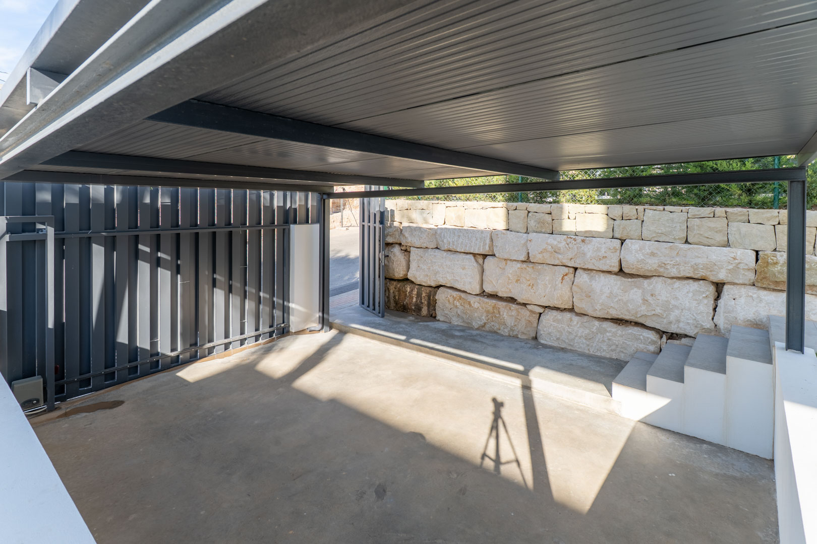 Privé overdekte parkeerplaats met een metalen constructie, betonnen vloer en een natuurstenen muur met aangrenzende trap naar de woning.