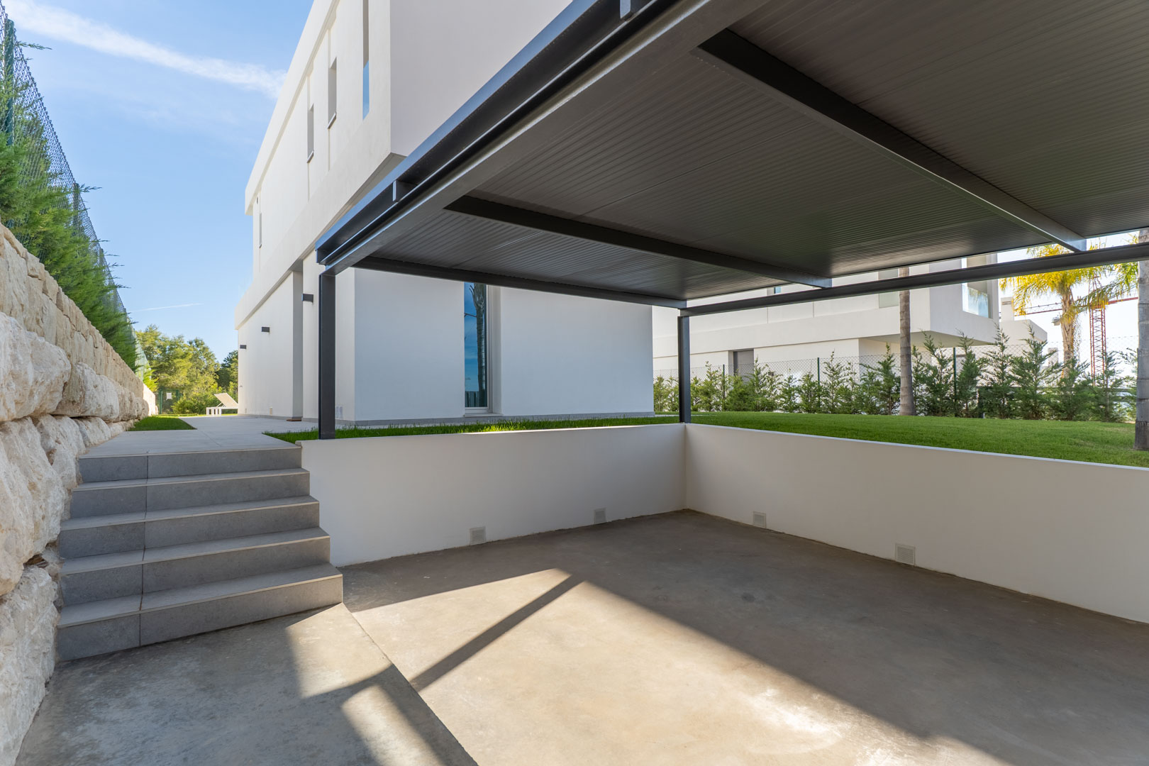 Privé parkeerplaats met een metalen carport, betonnen vloer en betegelde trappen die toegang bieden tot de tuin en de villa.
