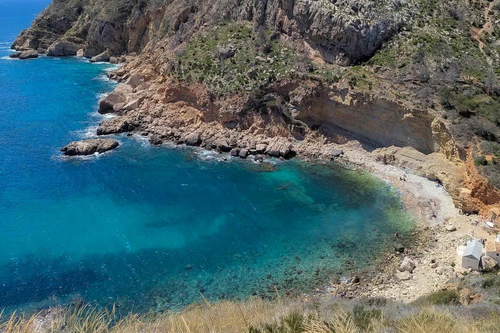 Luchtfoto van een natuurlijke baai in Moraira met kristalhelder turquoise water, steile kalkoeverwanden en een afgelegen kiezelstrand.