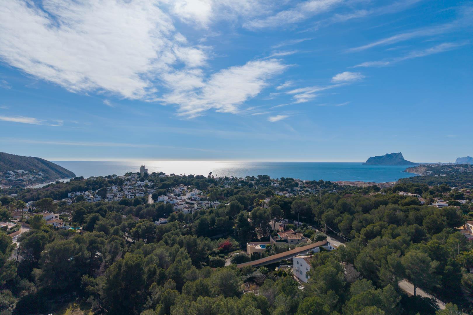 Panoramisch luchtfoto van de kustlijn van Moraira, met dichte dennenbossen, residentiële villa's en de Peñón de Ifach aan de horizon van de Middellandse Zee.