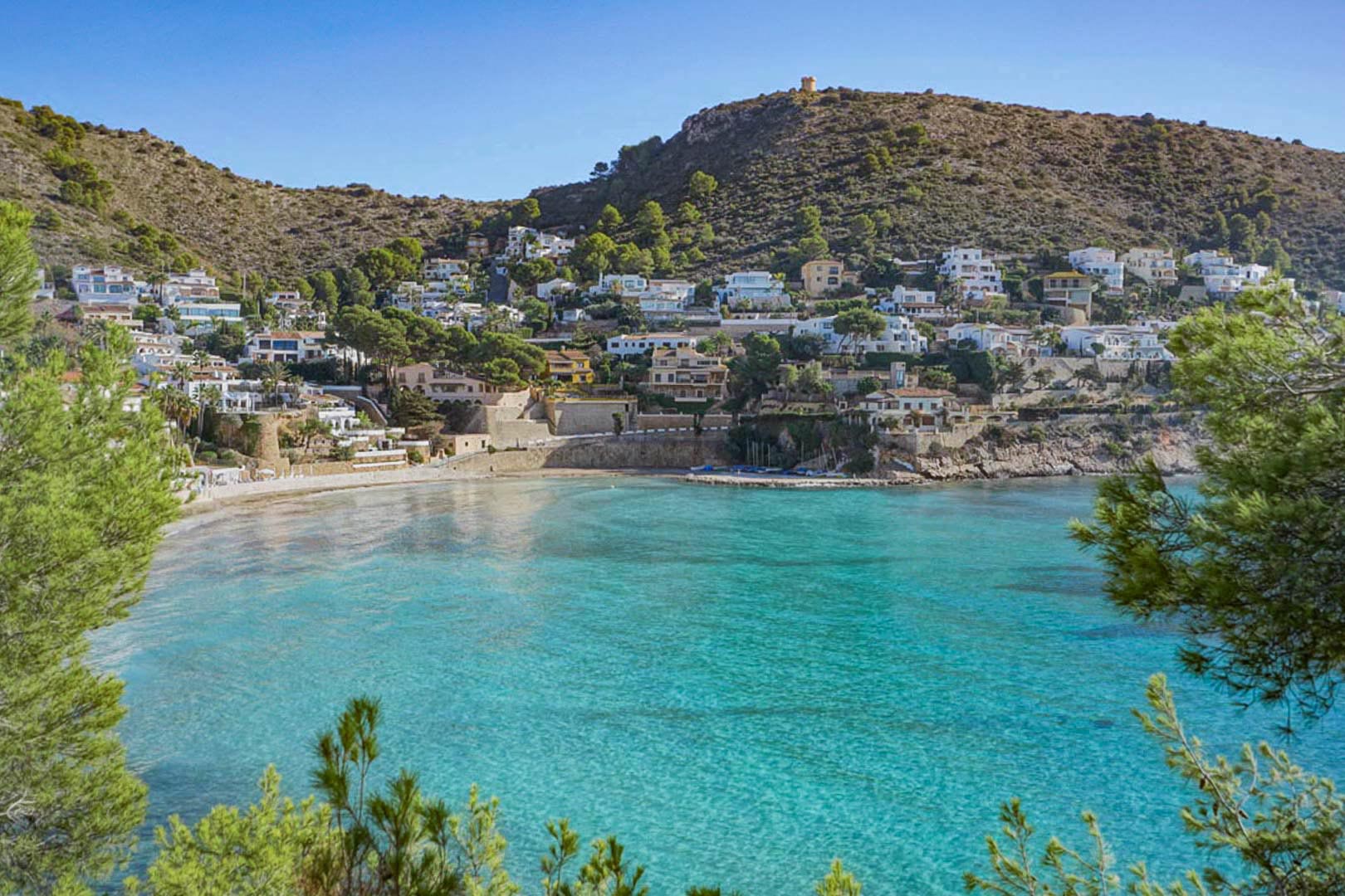 Panoramisch uitzicht op de baai van El Portet in Moraira, met kristalhelder water, een zandstrand en mediterrane villa's tegen de heuvel onder de historische uitkijktoren.