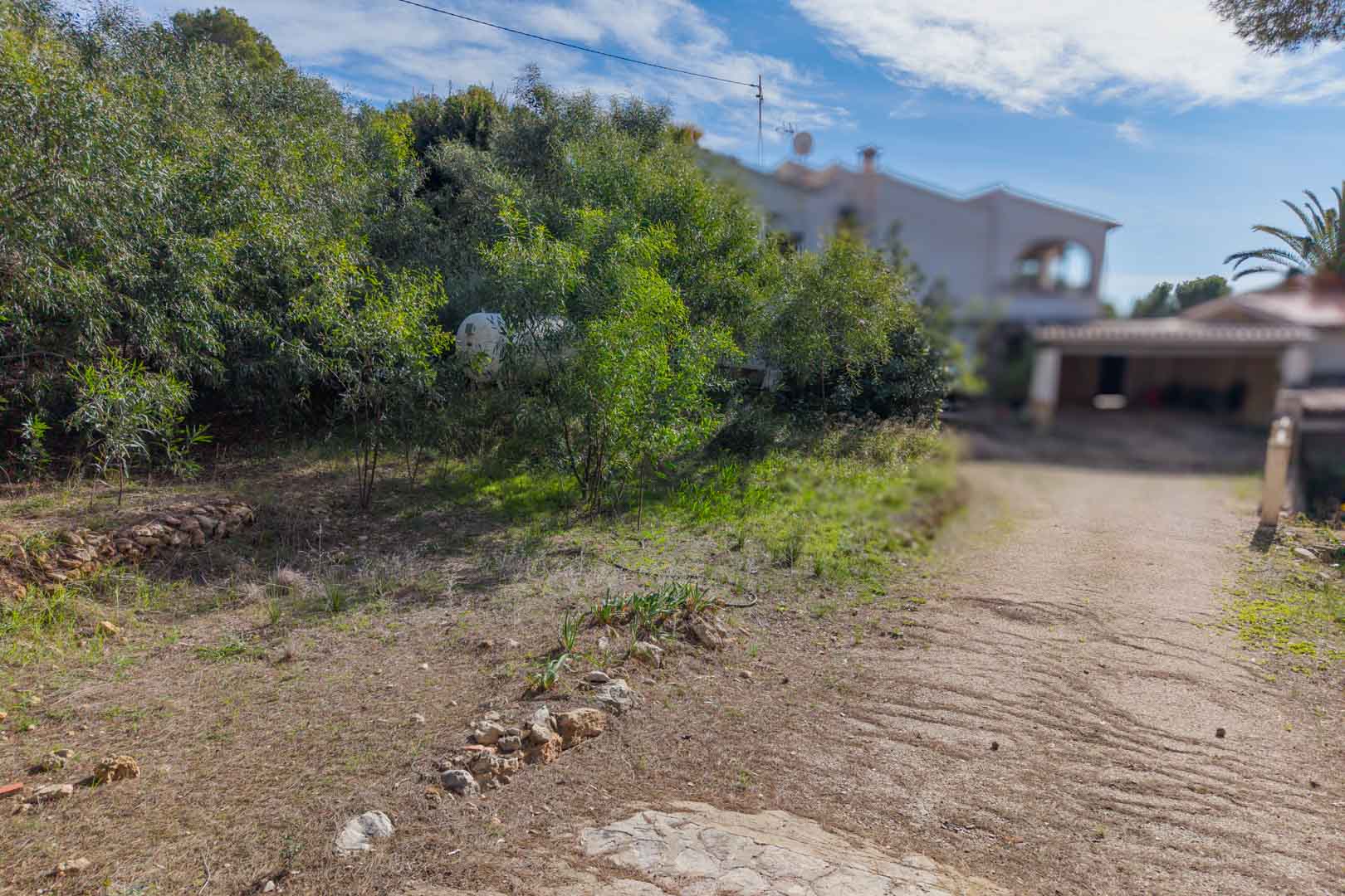 Toegangsweg naar het bouwperceel met natuurstenen randen en mediterrane struiken, met een bestaande villa op de achtergrond.