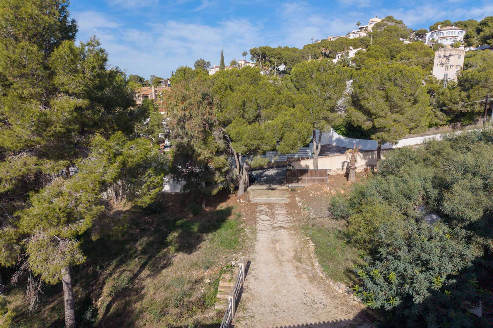 Verhoogd zicht op het bouwperceel met privé-toegangsweg, omgeven door volwassen pijnbomen en mediterrane beplanting in een gevestigde woonwijk.