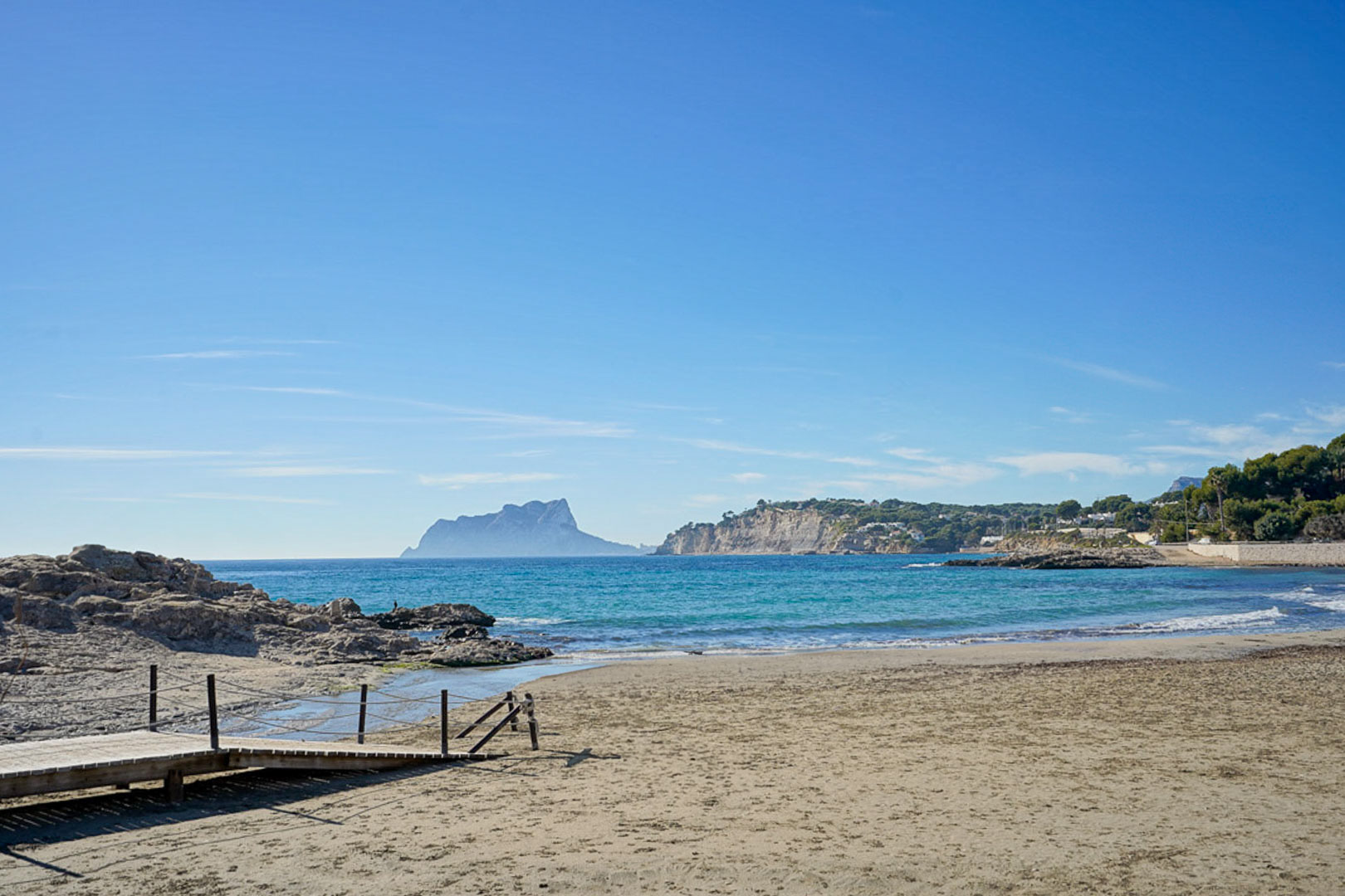 Zandstrand in Moraira met houten toegangspad, helderblauw zeewater en panoramisch uitzicht op de Peñón de Ifach in de verte.
