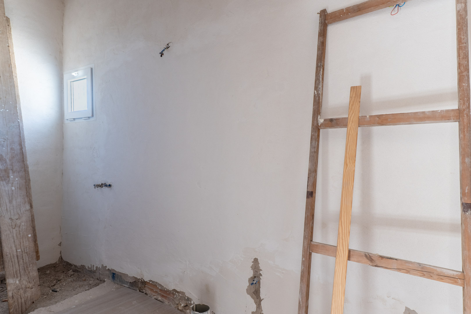 Binnenruimte in renovatiefase met vers gestuukte muren, een klein wit kozijn en voorbereide elektrische aansluitingen.