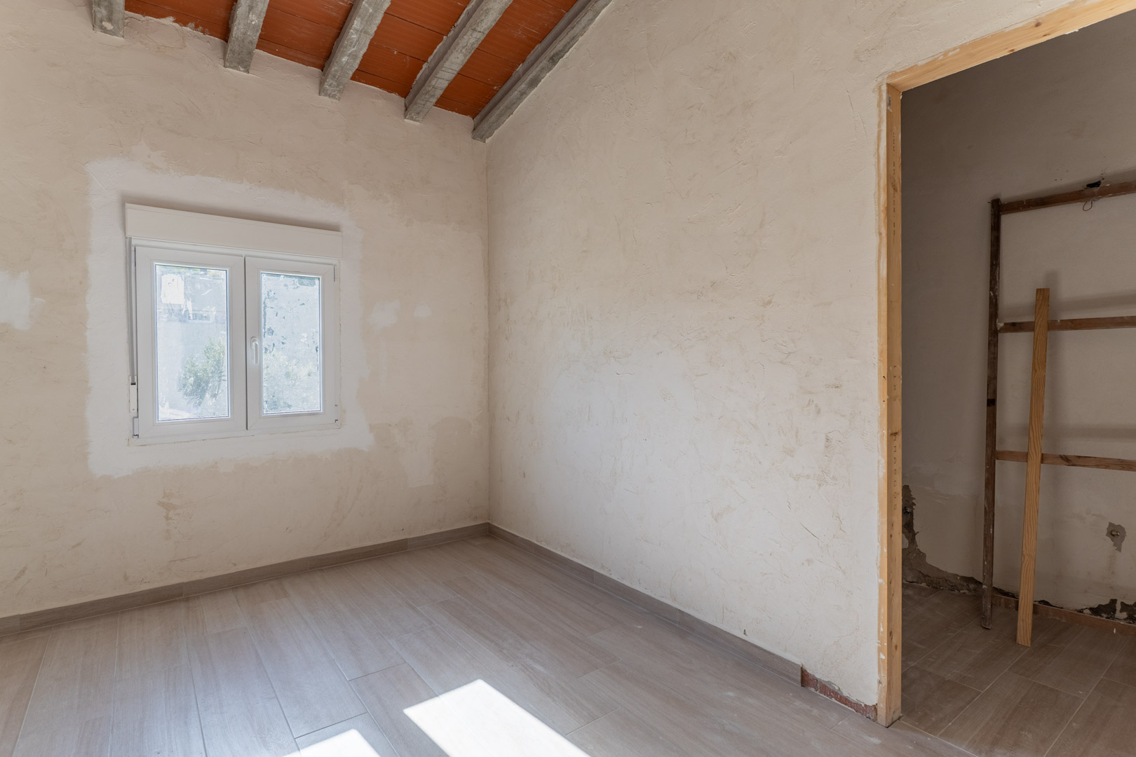 Gerenoveerde kamer met keramische vloertegels in houtlook, wit PVC-raam en een schuin plafond met zichtbare betonnen balken en terracotta stenen.