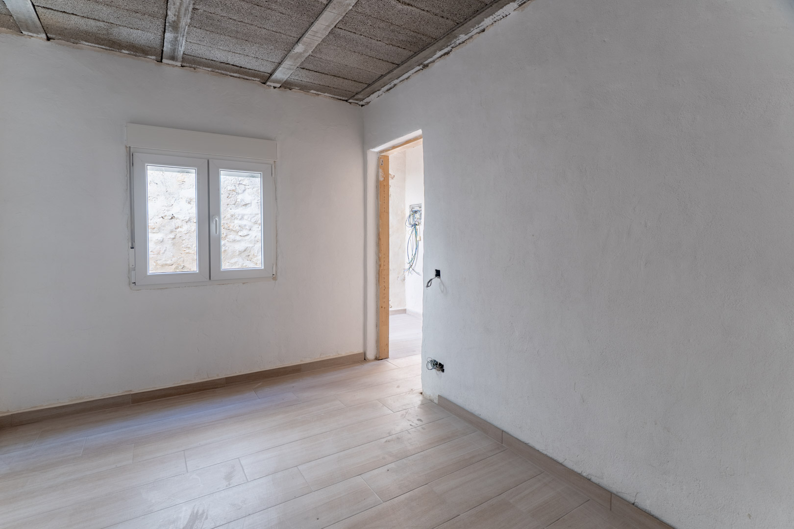 Gerenoveerde ruimte met keramische houtlook vloer, witte wanden en een nieuw PVC-raam. Het plafond toont de zichtbare betonnen balkenstructuur.