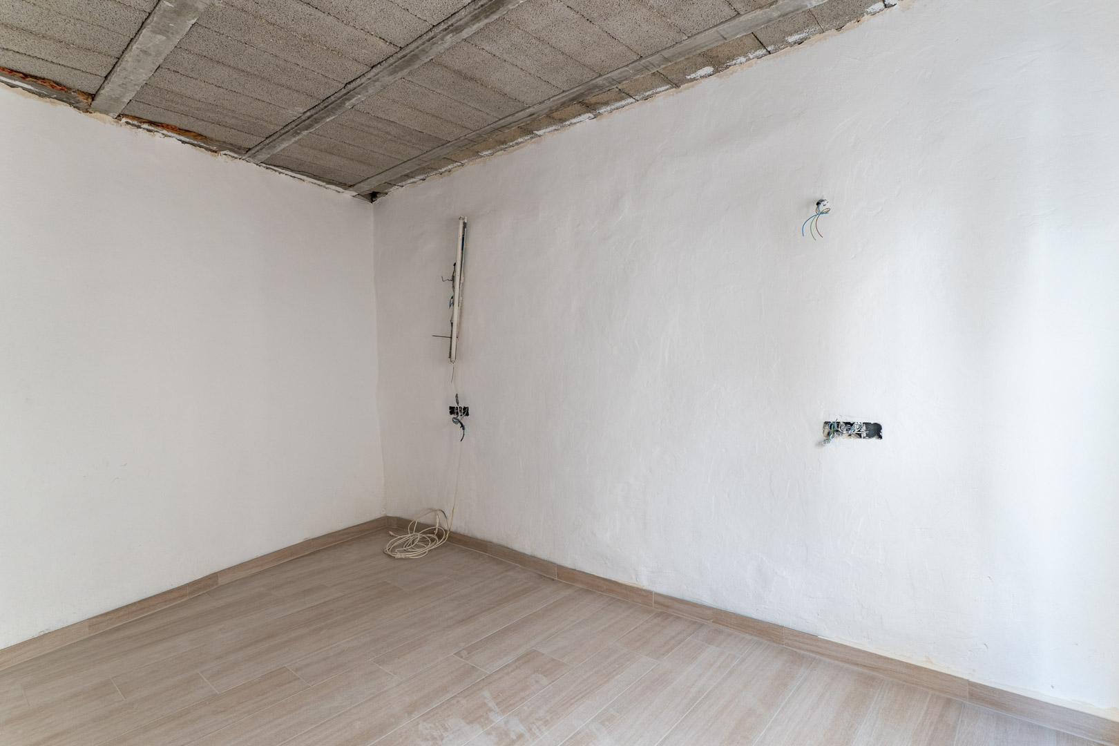 Te renoveren kamer met keramische vloertegels in houtlook, witte muren en een plafond van betonnen balken met zichtbare elektrische bedrading.