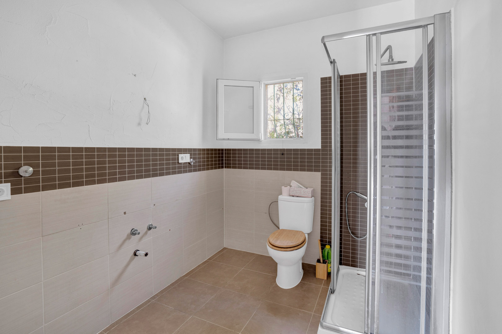 Badkamer met inloopdouche voorzien van glazen wand, bruine mozaïektegels en een toilet met houten zitting. De ruimte heeft neutrale wandtegels en een raam voor natuurlijke ventilatie.