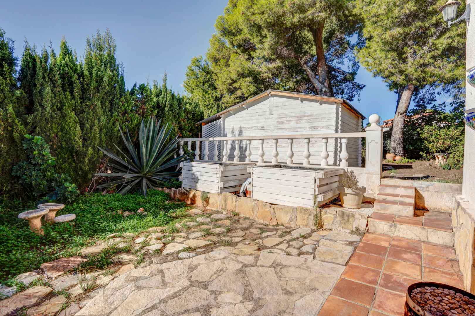 Buitenruimte met onregelmatige natuurstenen bestrating, een wit houten berging en mediterrane beplanting met pijnbomen en een grote agave.
