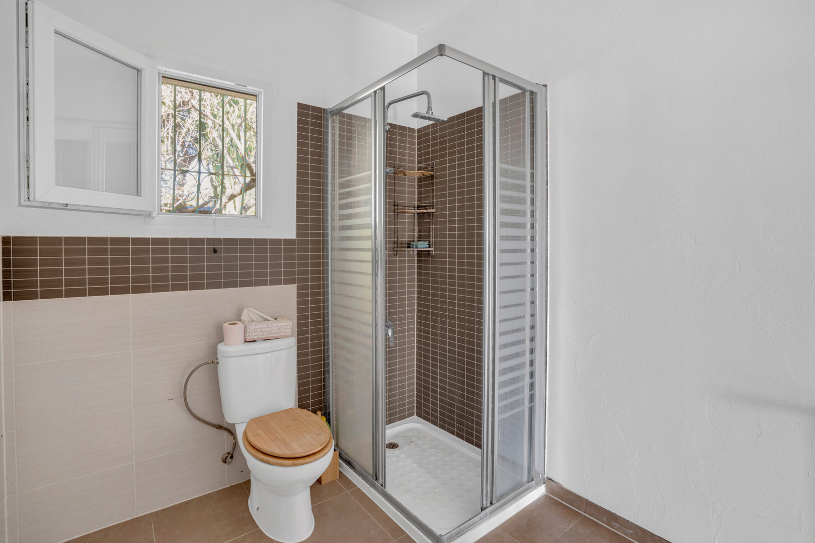 Functionele badkamer met een hoekdouche voorzien van bruine mozaïektegels, glazen wand, toilet met houten deksel en een raam voor natuurlijke ventilatie.