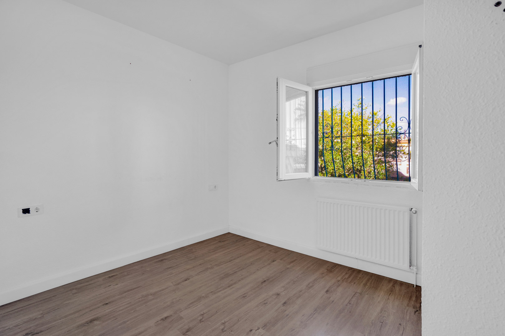 Lichte kamer met laminaatvloer in houtlook, witte muren en een raam met decoratieve tralies dat uitkijkt op de tuin, voorzien van een radiator.