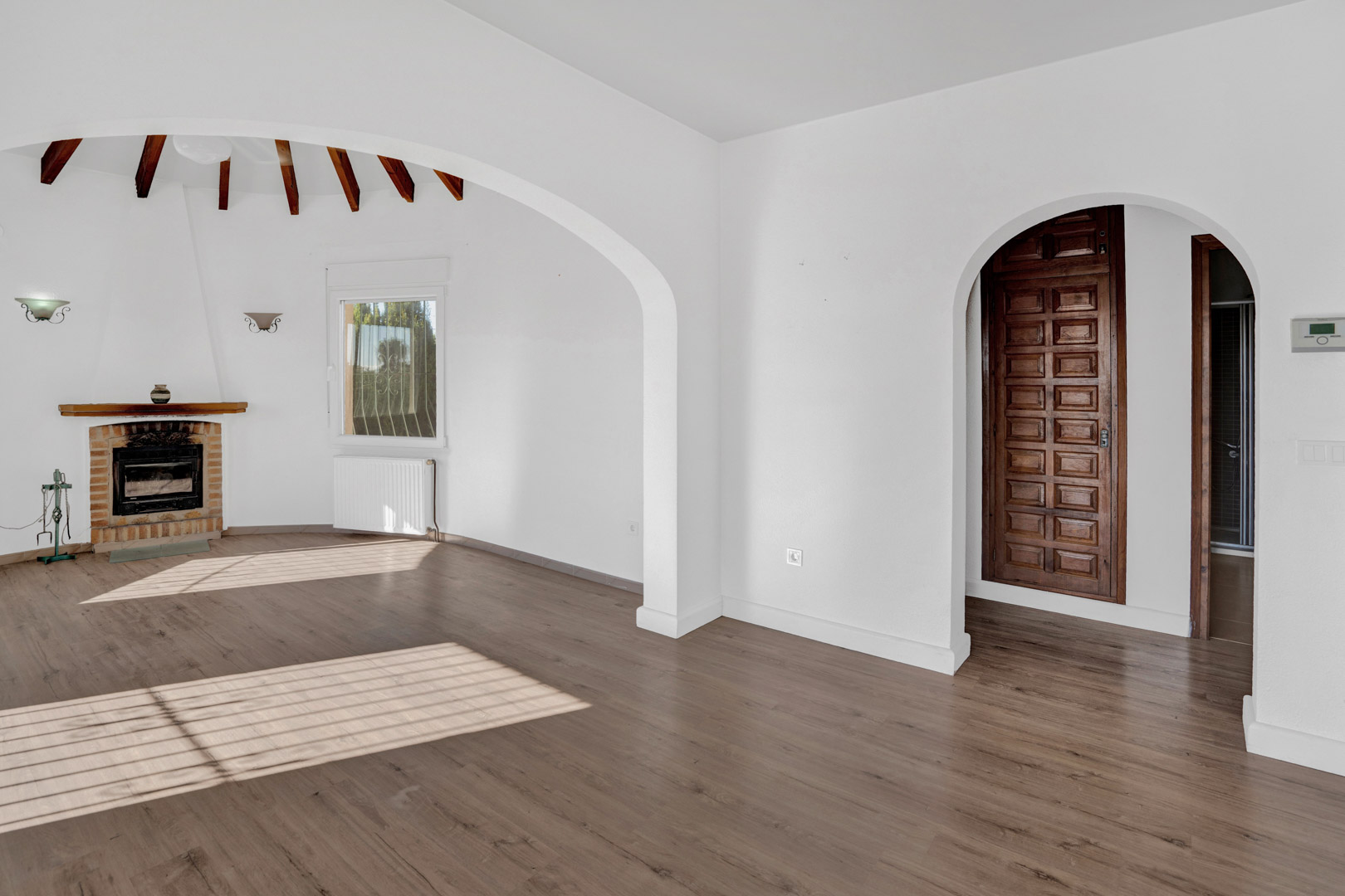 Lichte woonkamer met een bakstenen open haard, zichtbare houten balken en laminaatvloer. Voorzien van architectonische bogen, radiatorverwarming en een massief houten paneeldeur.