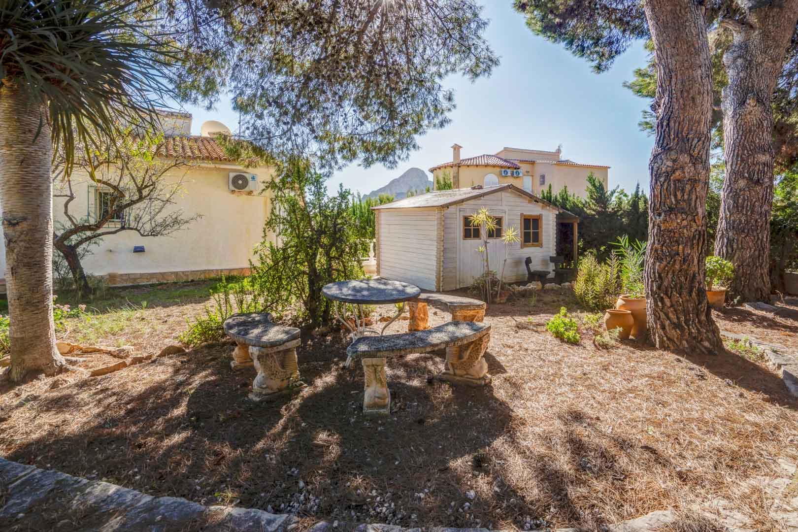 Mediterrane tuin met stenen zithoek onder volwassen bomen, een wit houten tuinhuis en uitzicht in de verte op de Peñón de Ifach.