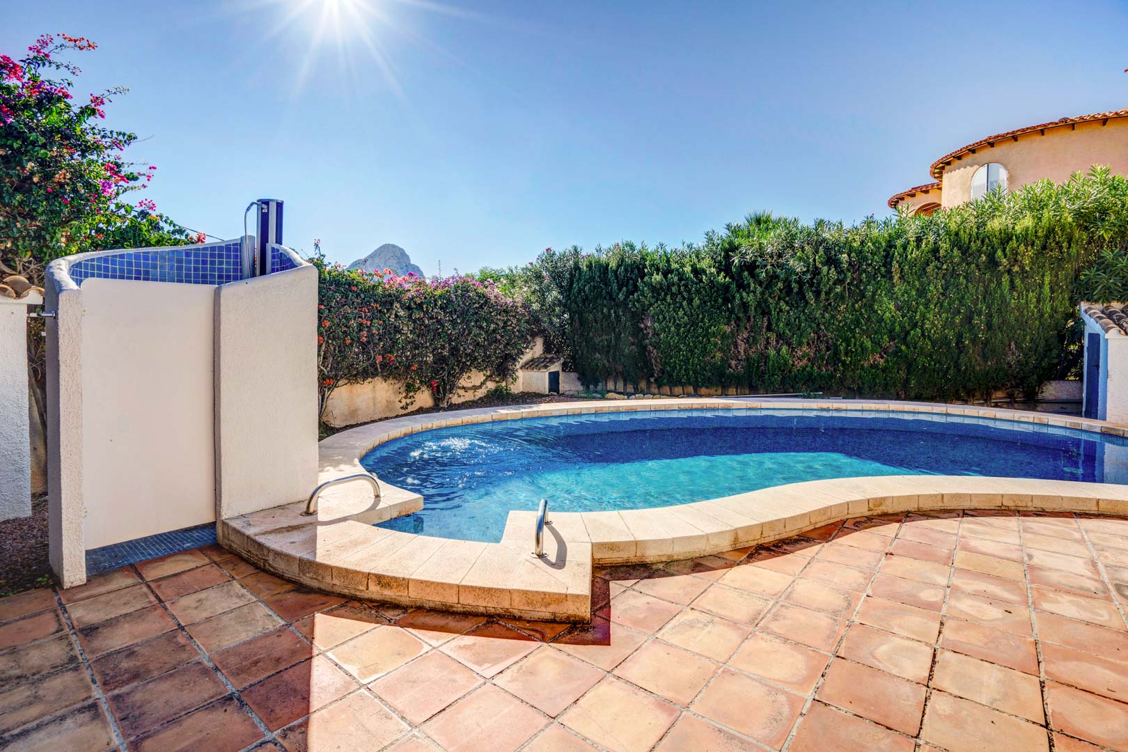 Nier-vormig privézwembad met een terracotta terras, buitendouche en uitzicht op de Peñón de Ifach achter volgroeide hagen die optimale privacy bieden.