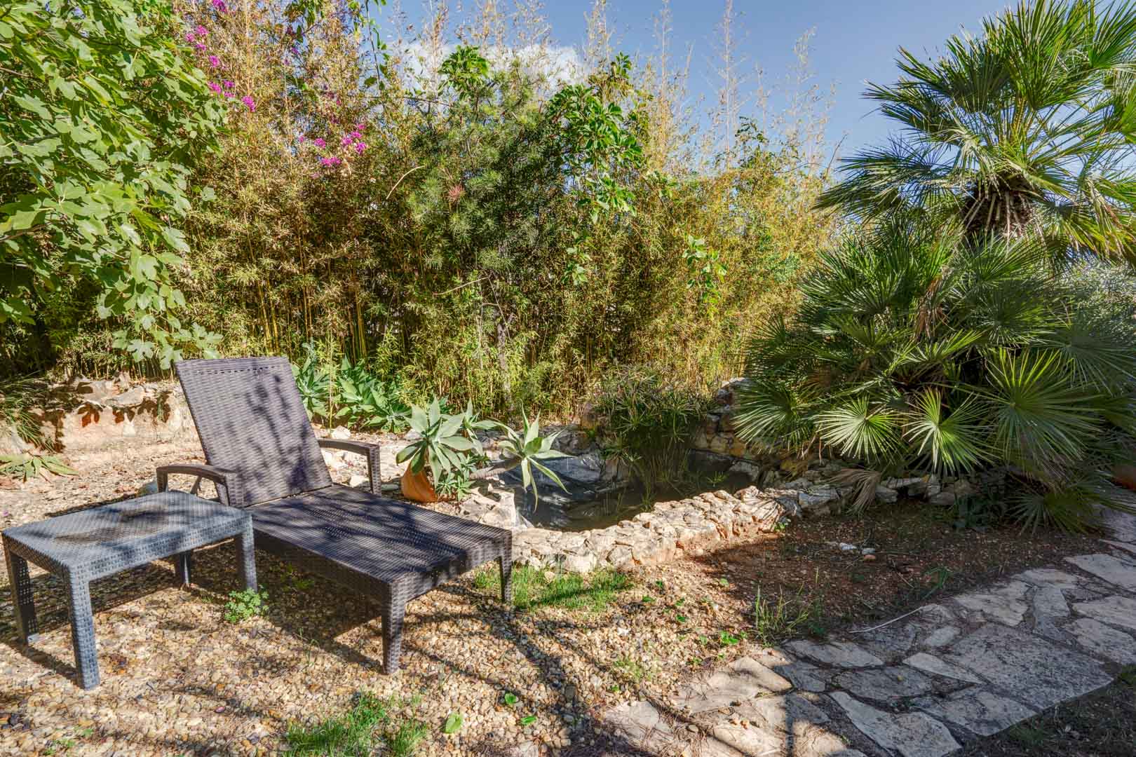 Ontspanningsruimte in de tuin met een rotan ligbed naast een stenen vijver, omgeven door volgroeide bamboe, palmbomen en mediterrane beplanting.