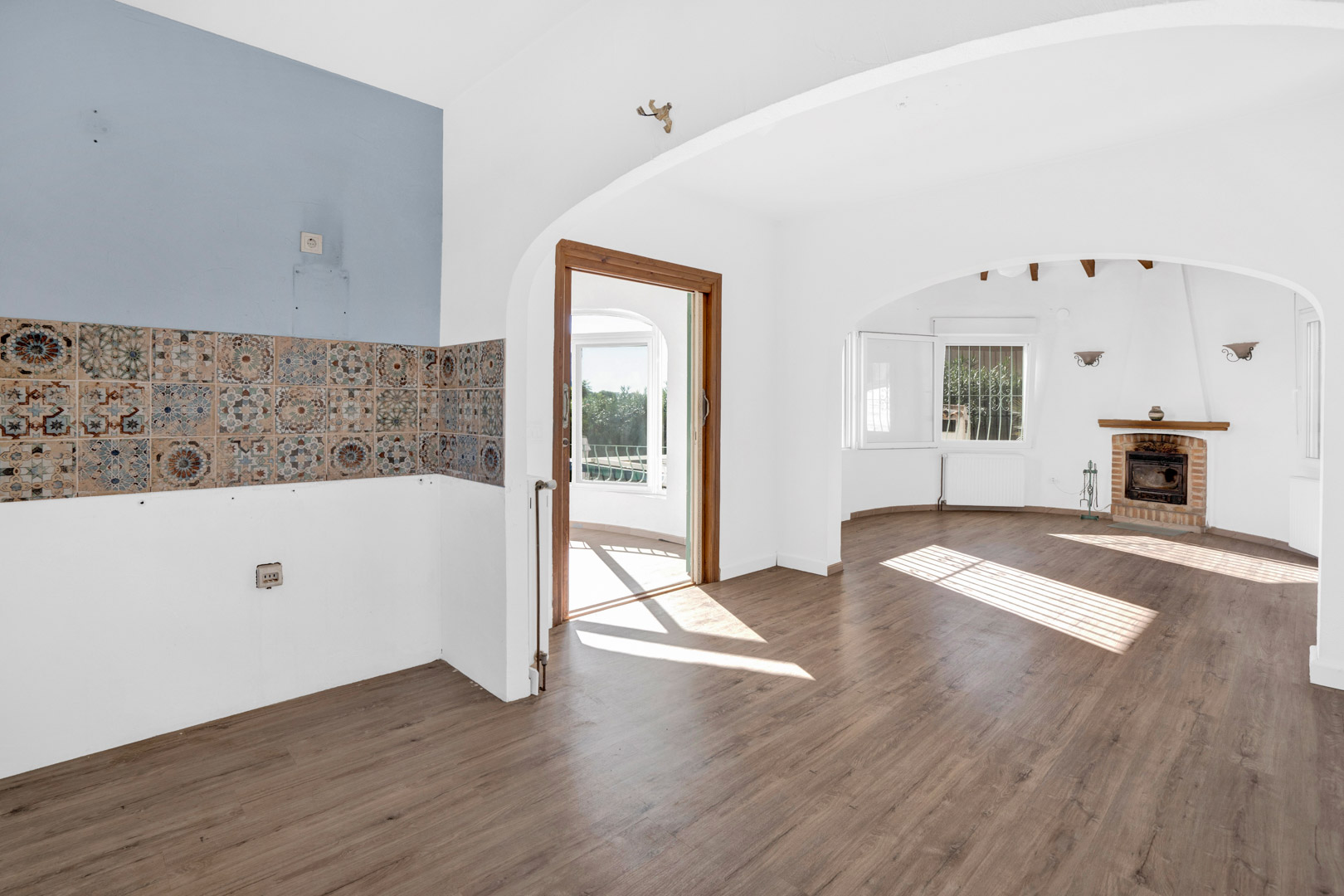 Open woonkamer met een bakstenen open haard, laminaatvloer en decoratieve keukentegels. Boogopeningen bieden toegang tot een lichte glazen serre.