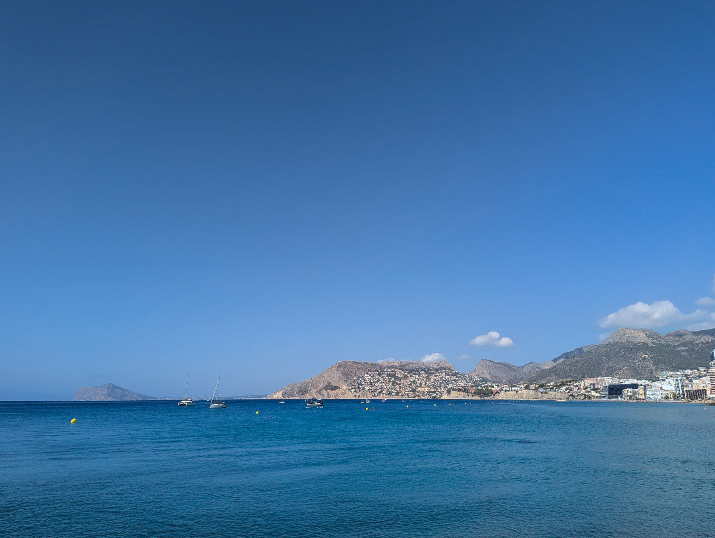 Panoramisch uitzicht op de Middellandse Zee en de kustlijn van Calpe met zeilboten en bergen aan de horizon onder een helderblauwe hemel.